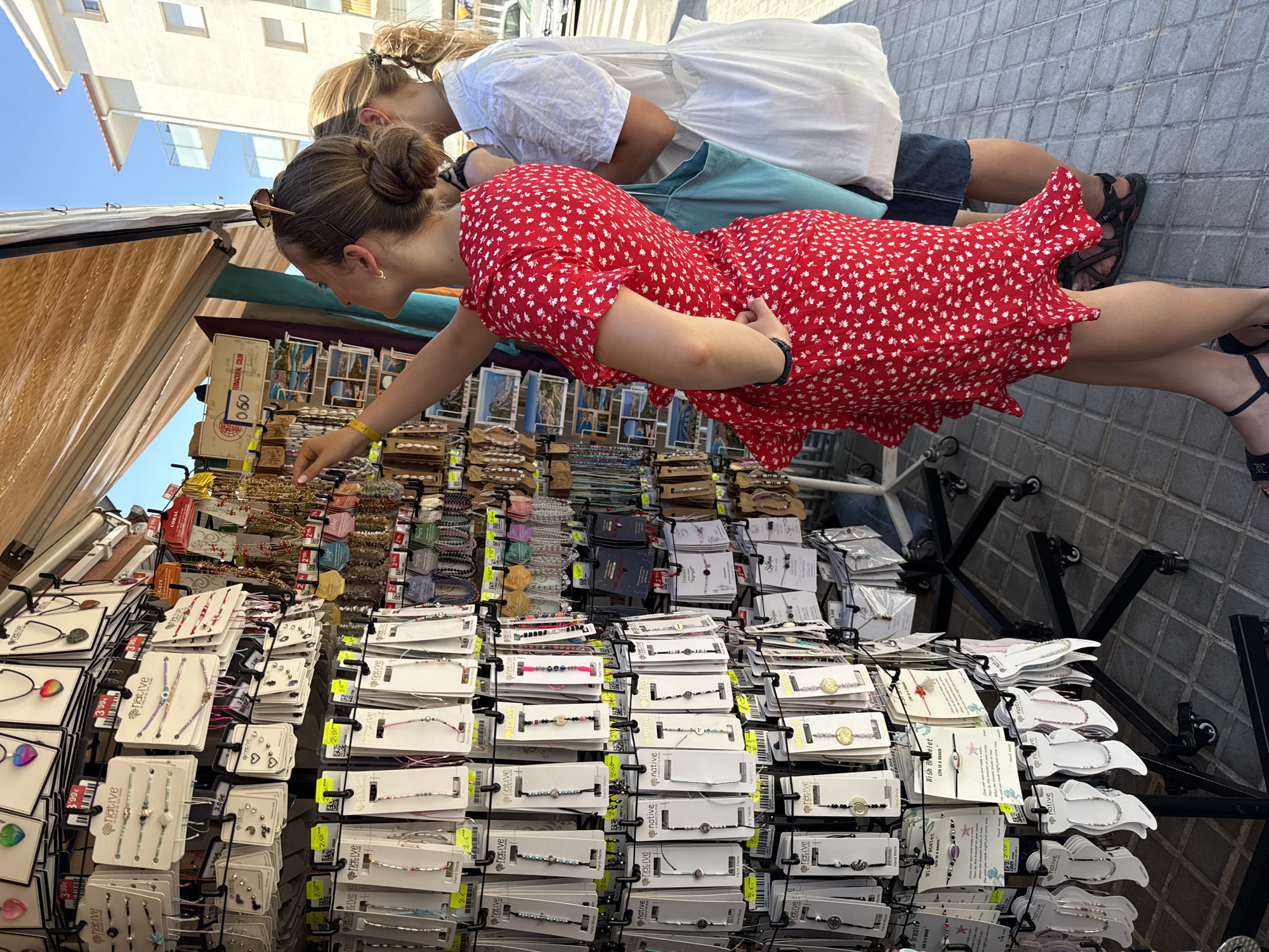 Two women are browsing a jewelry stand in Blanes, Spain, on a sunny day. The woman in the foreground, wearing a red floral dress and dark sandals, is reaching out to touch a piece of jewelry. She has sunglasses perched on her head. Behind her, another woman wearing a white shirt and dark shorts, holding a blue tote bag, looks on. The stand displays a variety of necklaces, bracelets, and earrings, many of which are packaged with price tags. The background shows buildings and a street, indicating an outdoor market or shop.