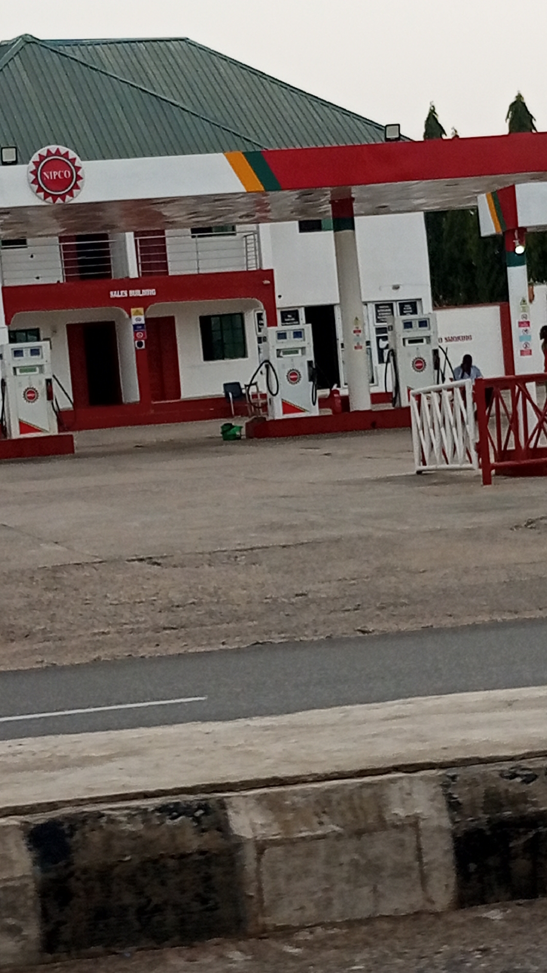 This image captures a NIPCO gas station in Adankolo, Nigeria, under what appears to be an overcast sky or diffused daylight.
The central subject is the gas station building and its forecourt. The building features a dark green, corrugated metal roof, white walls, and prominent red accents. A large, circular "NIPCO" logo, red with white text, is affixed to the upper left side of the building. Below the roofline, a decorative stripe in red, yellow, and green runs across the front of the structure. The words "SALES BUILDING" are clearly visible in white capital letters on a red band above the ground floor.
The forecourt is made of concrete and shows signs of wear. Several red and white gas pumps are arranged in front of the building, each displaying a circular stop sign symbol. Near one of the pumps, a black chair and a small green object are on the ground. To the right, white and red metal railings form a barrier, behind which a person wearing a light blue shirt can be seen. Another individual is partially visible further to the right. A partially visible sign on a white wall to the right of the pumps reads "NO SMOKING."
The foreground shows a concrete curb or barrier bordering an asphalt road, which has a white dashed line visible on its surface. In the background, above the gas station roof, several green trees are visible. The scene depicts an active but not busy gas station environment.