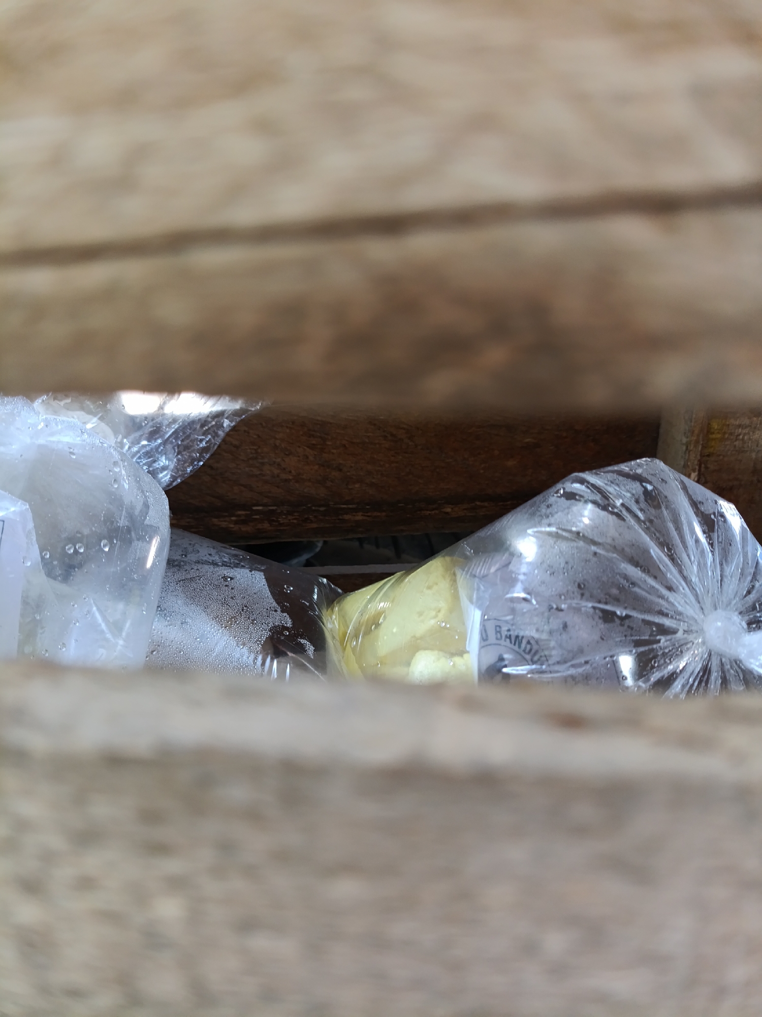 In Cariu, Indonesia, two plastic-wrapped items are visible through wooden slats. The item on the left appears to be a bag of ice or water, with condensation on the plastic. The item on the right is a yellow, solid object, also in plastic wrapping, with some condensation. A portion of text is visible on this second wrapper, reading "BANDUNG". The background consists of rough, wooden planks.