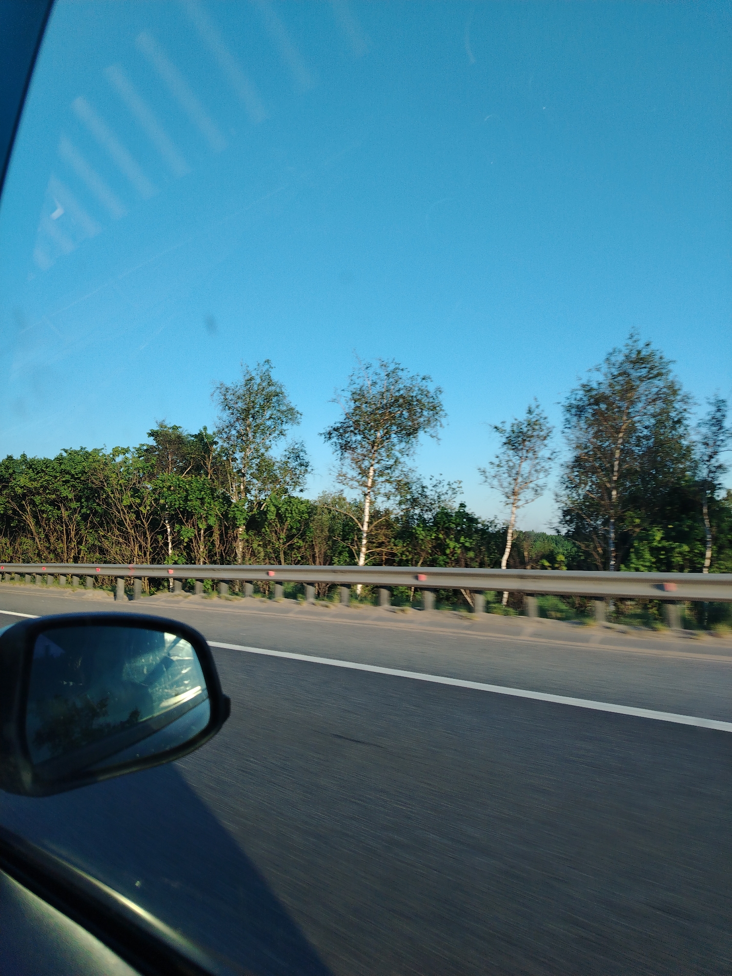The image depicts a view from a moving vehicle, likely a car, on a highway. A side mirror is visible in the foreground. A guardrail runs alongside the road, separating the highway from a wooded area with trees of varying heights. The sky above is clear and blue. The road surface is asphalt with a white lane marking. The scene does not clearly depict a city, but given the request for its location to be in Mitino, Russia, it is likely a highway or road in the outskirts or edges of that region.