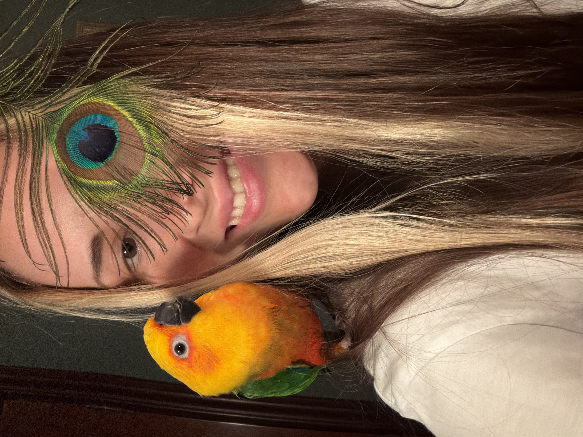 A close-up shot captures a woman and a parrot. The woman, with blonde and brown striped hair, is smiling and has a peacock feather partially obscuring her eye. The parrot, a Sun Conure, is perched on her left shoulder, its bright yellow and green plumage prominent. The background is dark, with a hint of a doorway on the left. The image does not provide any information about the city of Khimki, Russia.