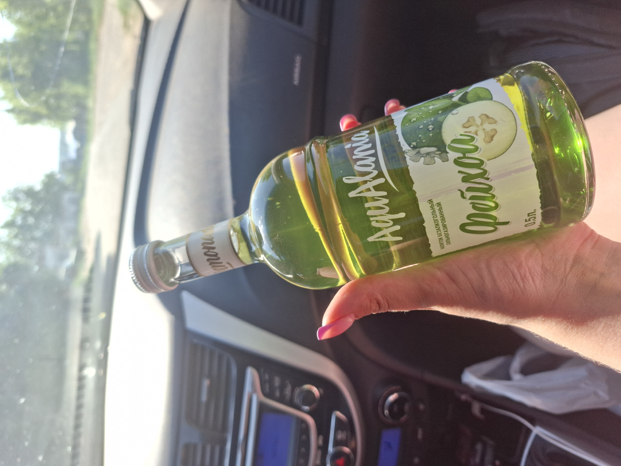 A hand with pink nail polish holds a bottle of "AquaAlania Фейхоа" sparkling drink inside a car. The drink is light green. The background shows the car's interior including the air vents and dashboard. The picture was taken in Chaikovsky, Russia.