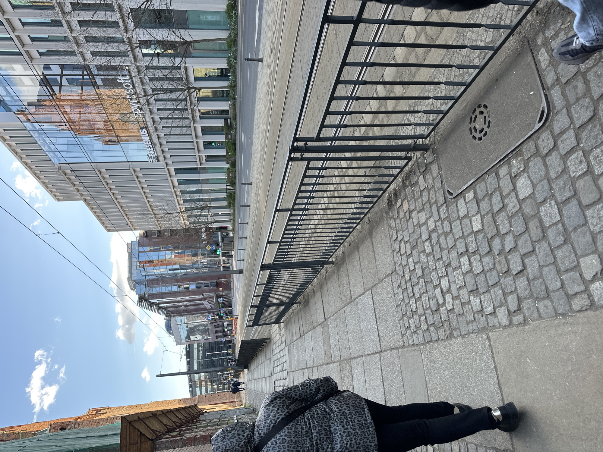The image depicts a street scene in Wrocław, Poland, with a modern building bearing the word "RENOMA" visible in the background. A pedestrian wearing a black and grey patterned jacket and black trousers is walking on the sidewalk. A black metal railing runs along the edge of the sidewalk. The street has tram tracks running down it, and there's a drain cover embedded in the cobblestone pavement. The sky is blue with some clouds. Other buildings are visible in the distance.