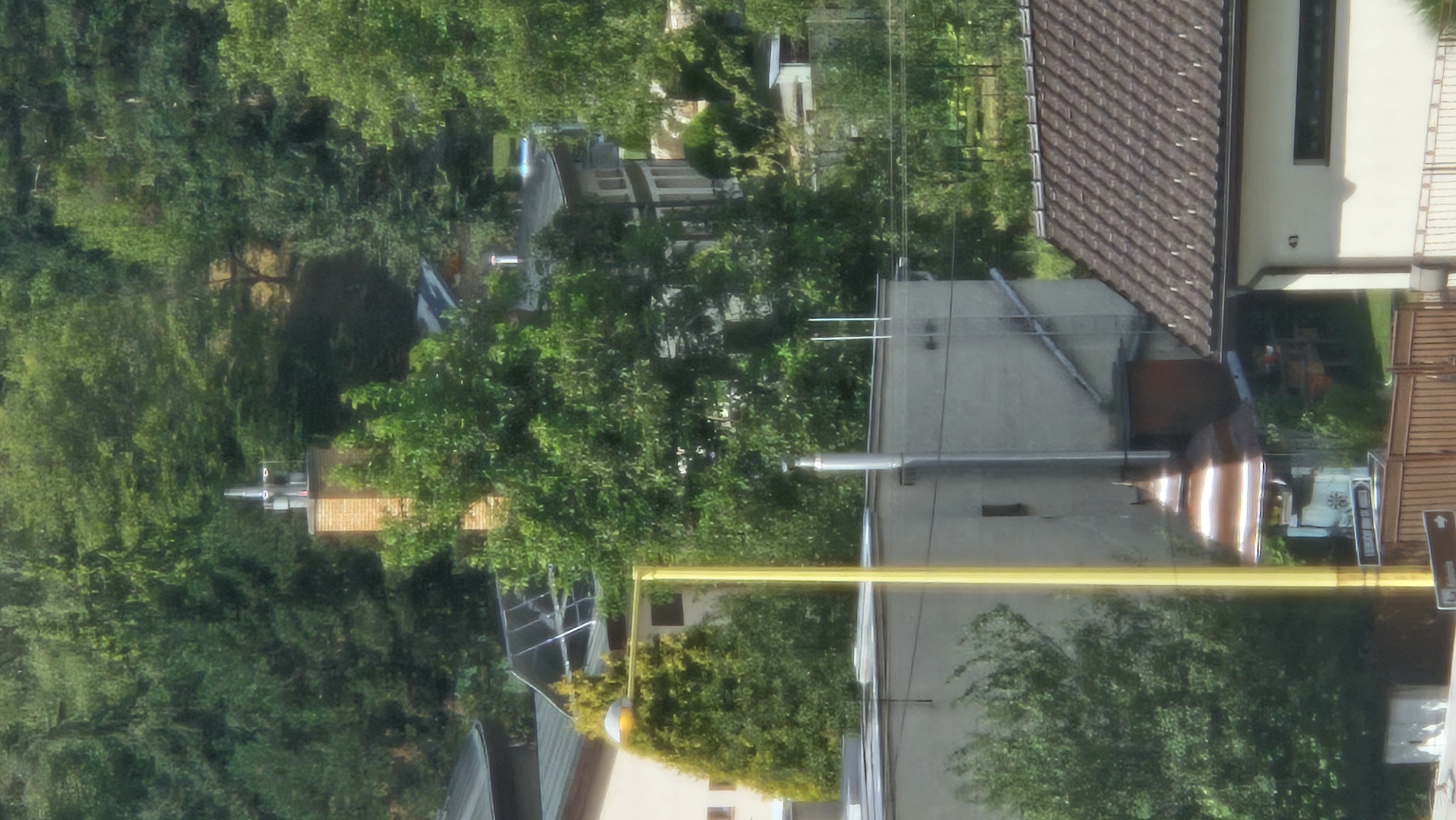 The image captures a dense, green treeline bordering a collection of residential buildings. In the foreground, a brown tiled roof of a building is visible, with the number '41' marked on its cream-colored facade. To the left of this building, a tall, light-colored structure with windows is partially obscured by foliage. A bright yellow lamppost stands prominently in front of this structure. Further to the left, a brick chimney with a metallic cap rises above the trees. The background is dominated by a verdant hill covered in numerous trees, suggesting a semi-rural or suburban setting. The scene appears to be a daylight shot with natural lighting. The architecture of the buildings suggests a European setting, possibly in the city of Prešov, Slovakia, given the context of the query, although no specific landmarks are identifiable to confirm this location.