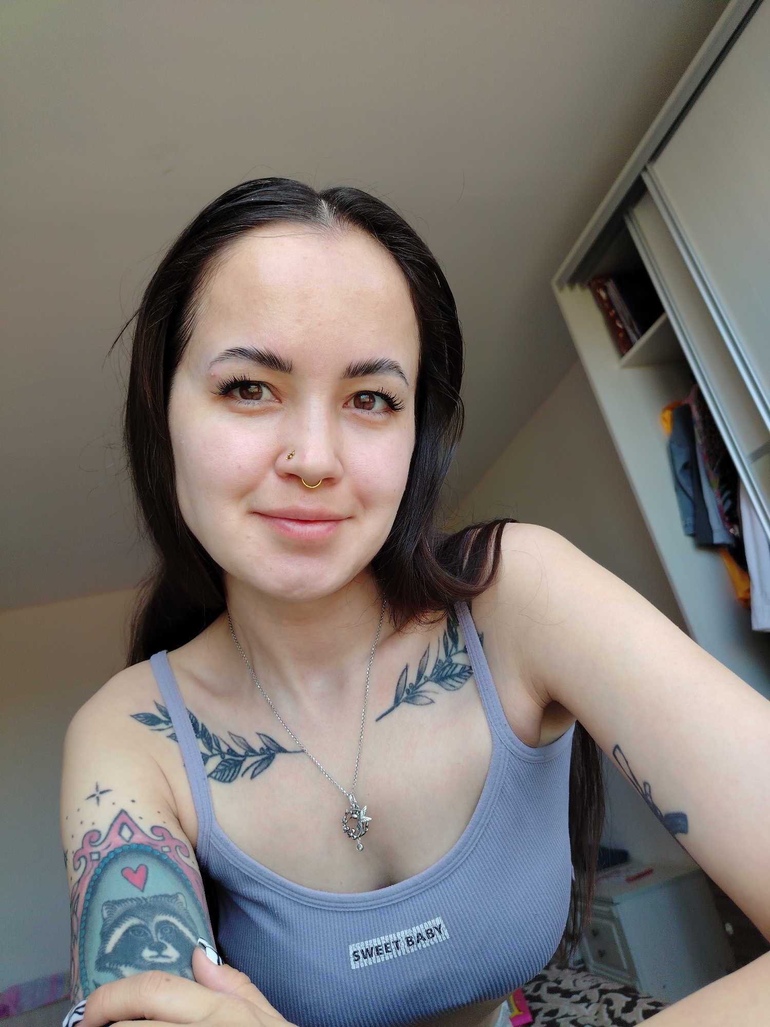 A selfie photograph depicts a young woman with dark hair and light skin. She is wearing a light purple tank top with the words "SWEET BABY" printed on the front. She has a nose ring and is wearing a silver necklace. The woman has tattoos on her arms and chest. A wardrobe is visible in the background. The location is not identifiable as Noviy, Russia, from the visual information.