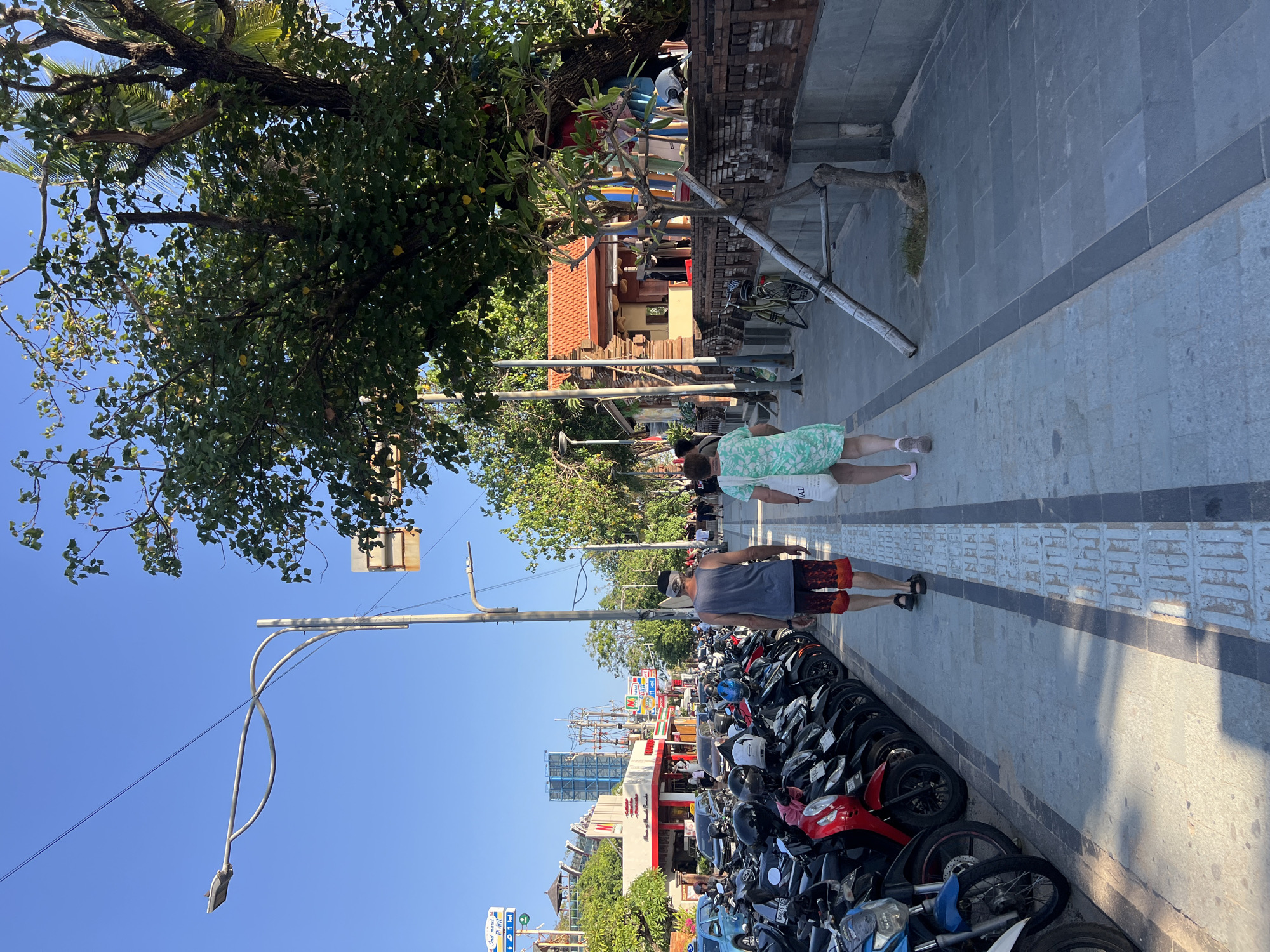 This image captures a scene on a street in Kuta, Indonesia, likely during the daytime. A paved sidewalk runs along the right side of the frame, bordered by a brick wall and lush trees. On the left, a row of parked motorbikes is visible, suggesting a busy area. Two individuals are walking away from the camera on the sidewalk. One, a male, wears a gray tank top and patterned red and black shorts. The other, a female, is in a green and white floral dress and carries a white tote bag. In the background, buildings, more motorbikes, cars, and electrical poles line the street, under a clear blue sky. The overall atmosphere appears to be that of a typical commercial street in a tropical urban environment.
