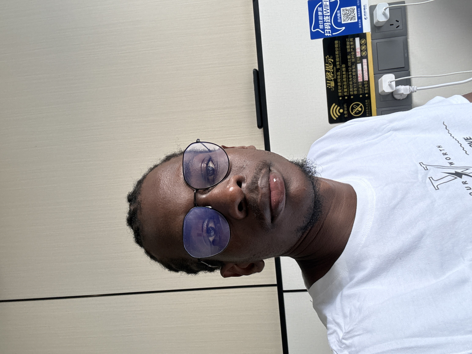 A man with dark skin and braided hair is shown in a close-up, selfie-style shot. He is wearing round, wire-rimmed glasses with a blue tint and a white t-shirt. The background consists of a textured, light-colored wall and a section of electrical outlets with plugs. A black object, possibly a power strip or a small appliance, is mounted horizontally on the wall above the outlets. Two signs are visible on the wall near the outlets. One sign is black with yellow text and a WiFi symbol, containing information in Chinese. The other sign is blue and white with Chinese text and a QR code, advertising WiFi services. The lighting appears to be artificial and bright, creating some glare on the man's glasses and skin. The environment suggests an indoor location, possibly a hotel room or a public space with charging stations. The Yuzhong District, China, is not explicitly identifiable from the visual cues within the image.