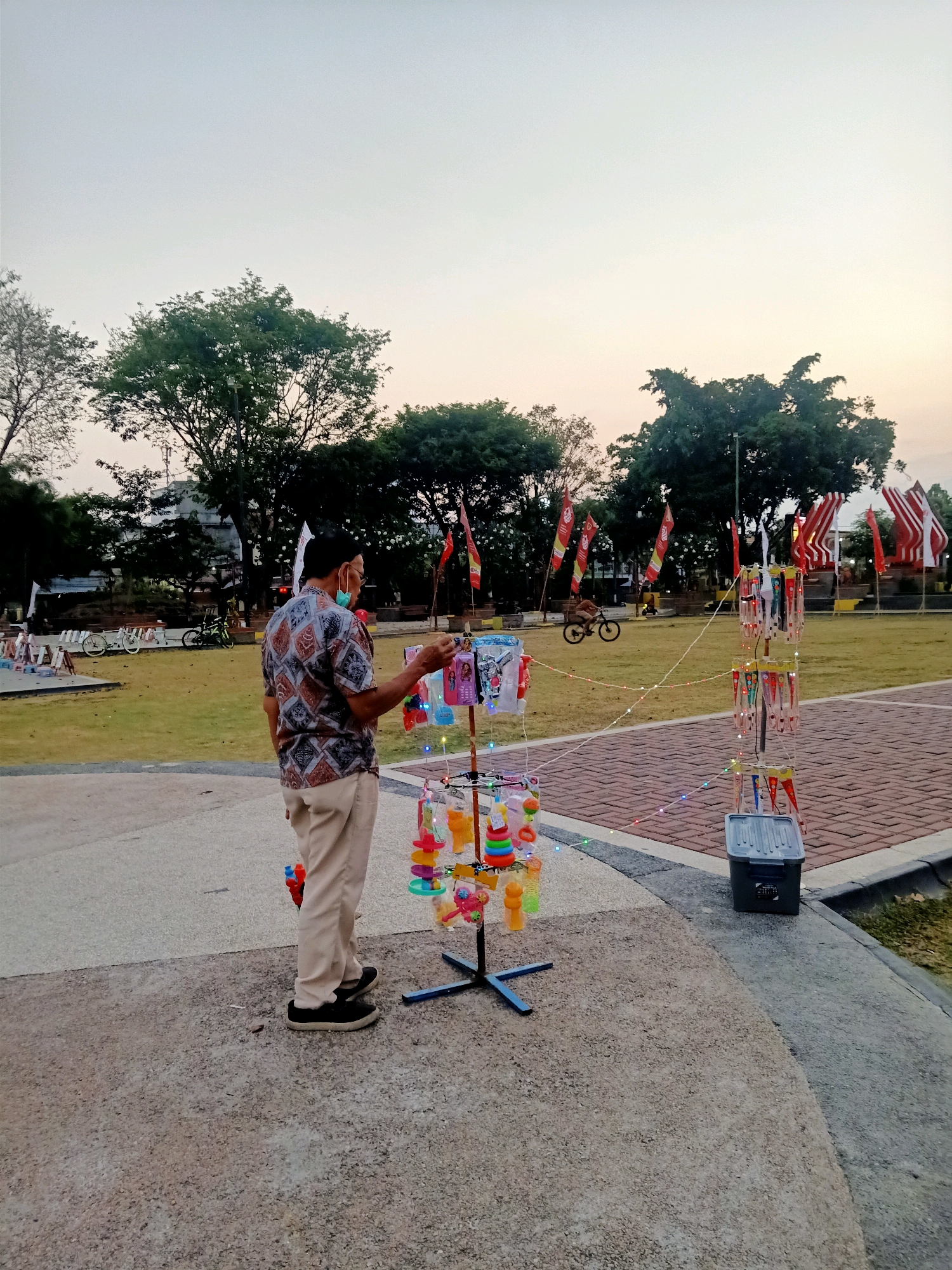 This is a medium shot of a man selling toys from a stand in an outdoor park area in Klaten, Indonesia, during the late afternoon or early evening. The man, wearing a patterned short-sleeved shirt, light-colored pants, black shoes, and a light blue face mask, stands behind a rotating display rack filled with colorful toys, including what appear to be plastic whistles and other small novelty items. The display is illuminated by small, multi-colored string lights. In the background, a paved walkway separates the man from a grassy lawn where several red and white flags on poles are visible, along with a monument or structure. Two bicycles are parked to the left, and a child is riding a bicycle on the lawn in the mid-ground. The sky is pale, suggesting either dawn or dusk. The overall environment appears to be a public park or recreational area.