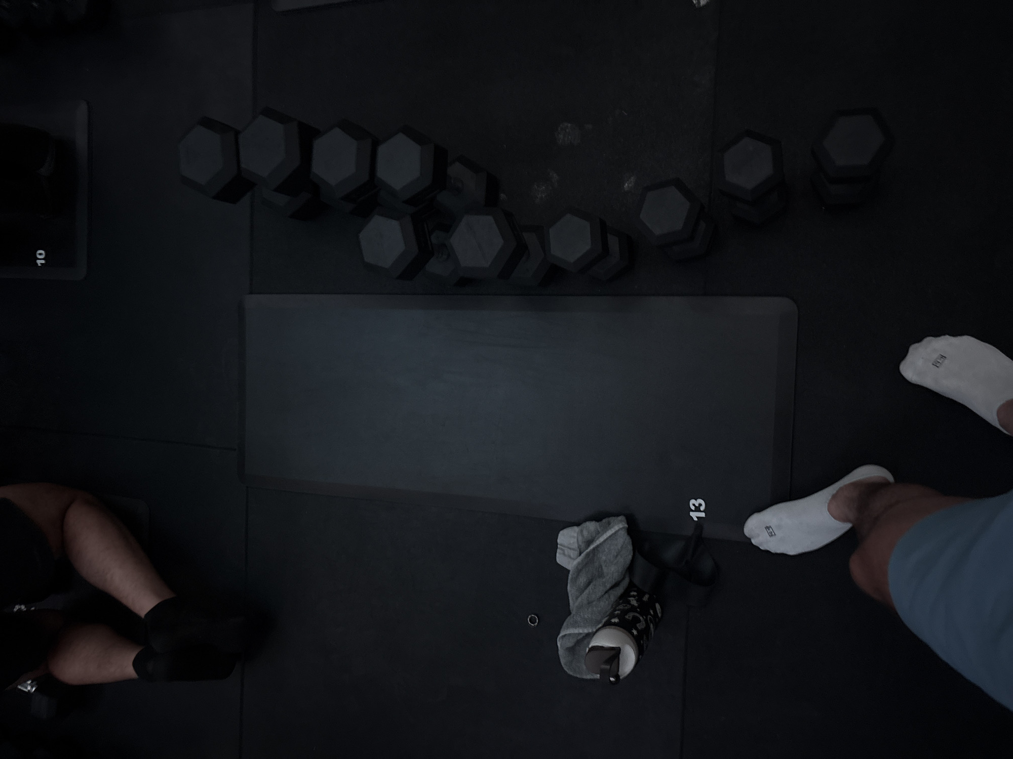 A dimly lit indoor workout area is observed from an elevated perspective. Various hexagonal-shaped dumbbells are arranged in stacks on the floor, with numbers '10' and '13' printed on separate square platforms. A dark workout mat occupies the center of the scene. A person wearing black socks kneels on the left side. Another individual wearing white socks and blue clothing is visible on the right side. A water bottle and a gray towel lie on the floor near the number '13' platform. The location is San Bernardino Tlaxcalancingo, Mexico.