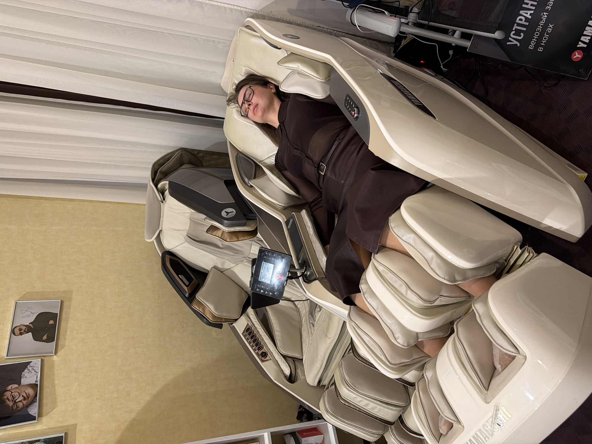 A woman with glasses lies in a Yamaha massage chair in a room in Izhevsk, Russia. She wears a brown dress and has dark hair. The massage chair is beige and has multiple pads for legs and arms. A control panel is visible near the woman's hand. The background includes a wall with framed pictures, curtains, and a black board with Russian text. The floor is brown.