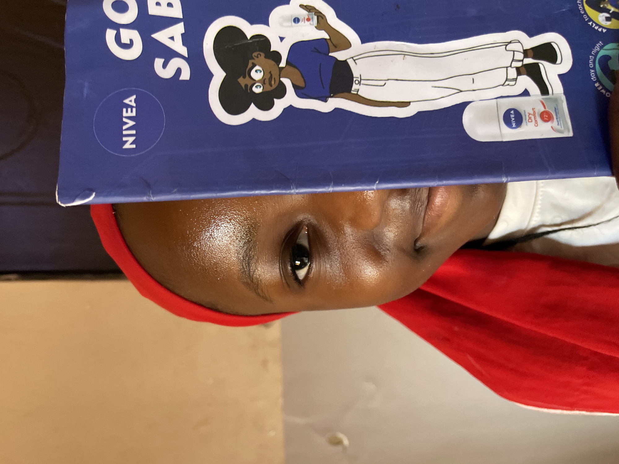 A close-up, top-down image features a person, likely in Jos, Nigeria, partially obscured by a blue book or folder. The person has dark skin, and only their forehead, nose, and one eye are fully visible, looking upwards with a neutral expression. They are wearing a red head covering or garment.
Dominating the foreground is a blue book or folder, positioned directly above the person's face. It features prominent white text, including "NIVEA" within a circular logo, and partial words "GO" and "SAB" (potentially part of "GOOD SABAH" or "GOOD SABAHI"). A cartoon illustration of a dark-skinned woman with dark curly hair is prominently displayed on the blue surface, holding a small NIVEA product in her hand. Below this illustration, a white NIVEA Dry Comfort deodorant stick is placed horizontally on the book, clearly showing "NIVEA", "Dry Comfort", and "72h". Other faint text on the blue surface includes phrases like "APPLY to each", "day and night", and "POWER over and might".
The setting appears to be indoors, with a plain, light-colored background suggesting a wall or floor. The lighting is bright and even, highlighting the person's skin and the details on the book. The activity seems to involve the person engaging with or being photographed in relation to the NIVEA promotional material.