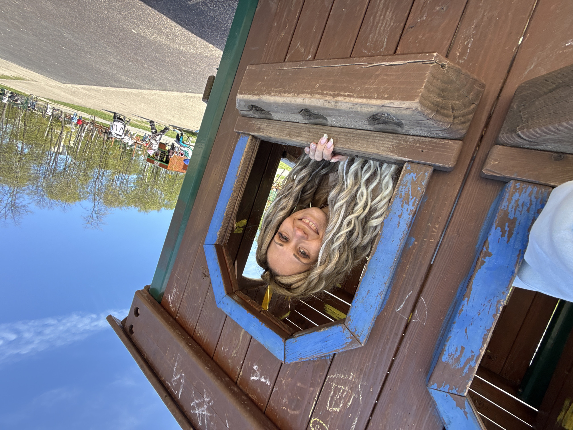 A woman with blonde dreadlocks is smiling through a blue-framed opening in a weathered wooden structure. The structure appears to be part of a playset or small building. The background shows a blue sky and trees, with what looks like a road and some kind of market or fair setup in the distance. The image is angled as if the camera is tilted. The location is Заволжье, Russia.