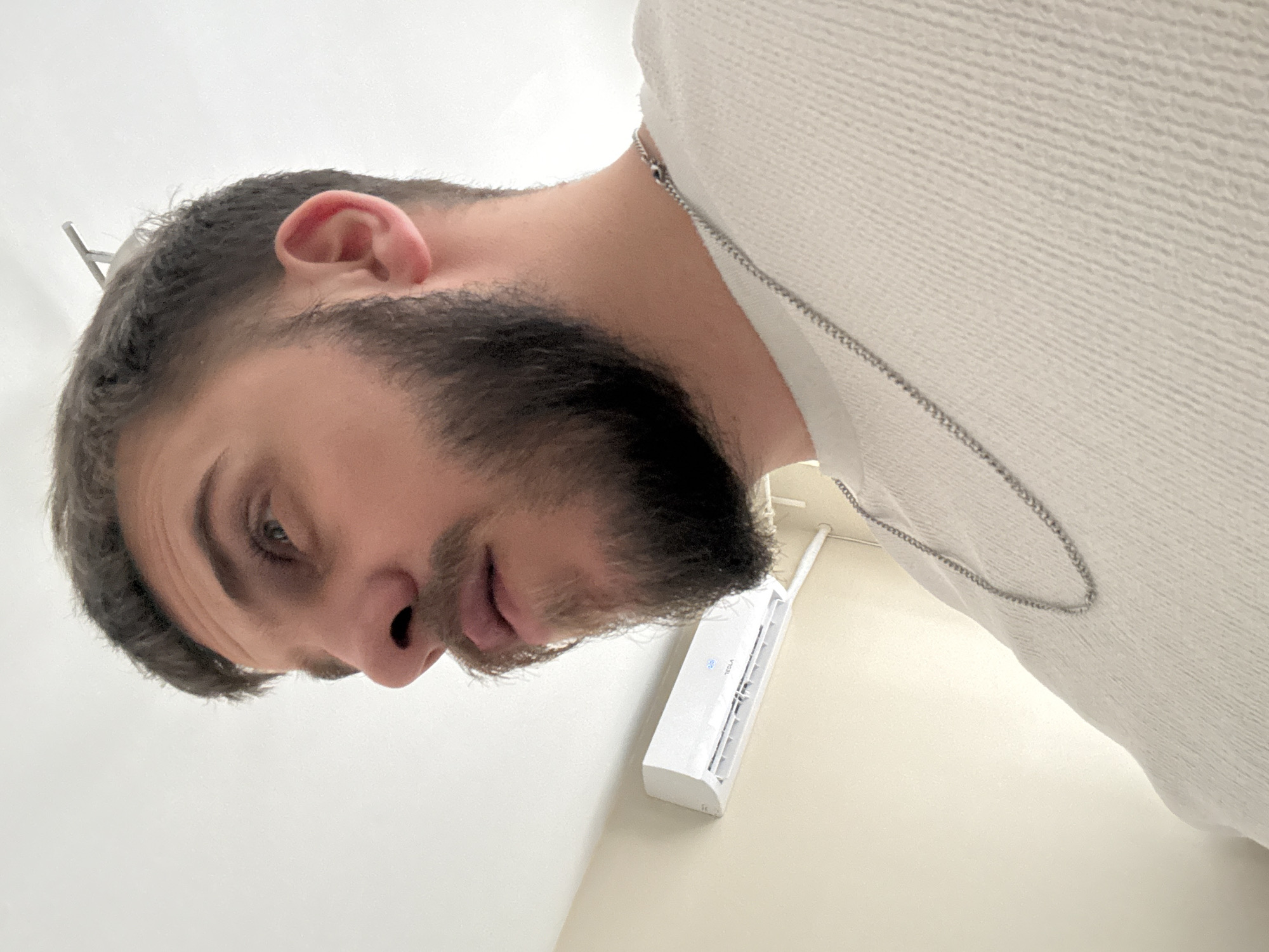 A close-up, upside-down selfie of a man with graying dark hair and a full, dark beard. He is wearing a white sweater with a silver chain. He is indoors with white walls. A white TESLA mini split air conditioner is mounted on the wall. There is no indication in the image of the location of the city of Sofia, Bulgaria.