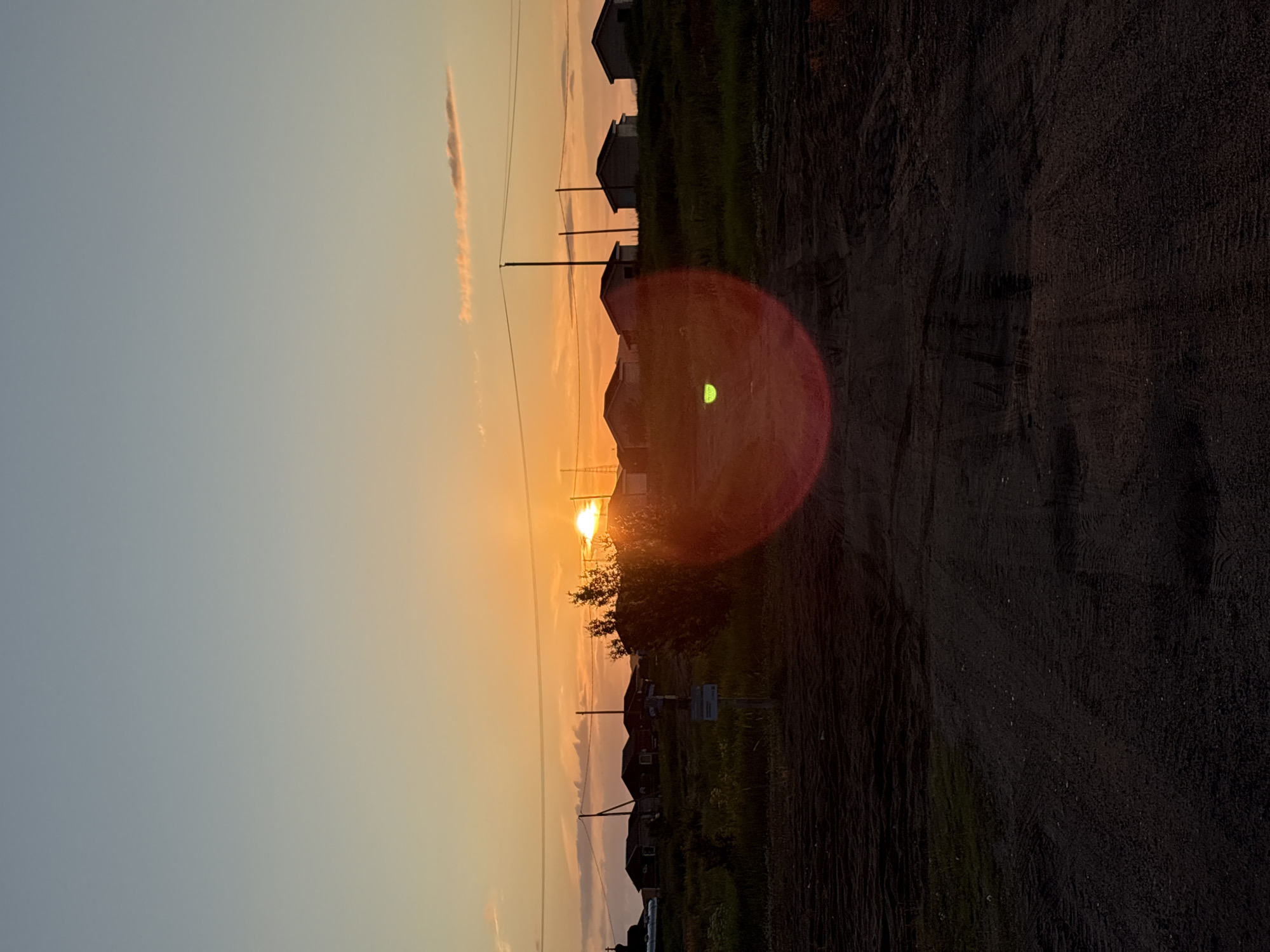 The image depicts a rural landscape at sunset in Teriberka, Russia. The sun, a bright orange orb, is low on the horizon, casting a warm glow across the sky which transitions from a pale blue to a soft orange. Silhouetted against the vibrant sky are several small, dark-colored houses, their roofs and walls indistinct. Power lines crisscross the scene, supported by tall, slender poles. In the foreground, a dirt road or path leads towards the houses, its surface appearing rough and uneven. Vegetation, likely grass and shrubs, is visible on the embankment where the houses are situated. A notable lens flare, a circular reddish-orange glow, is prominent in the lower center of the image, obscuring some of the foreground details. The overall atmosphere is one of quietude and the fading light of day.