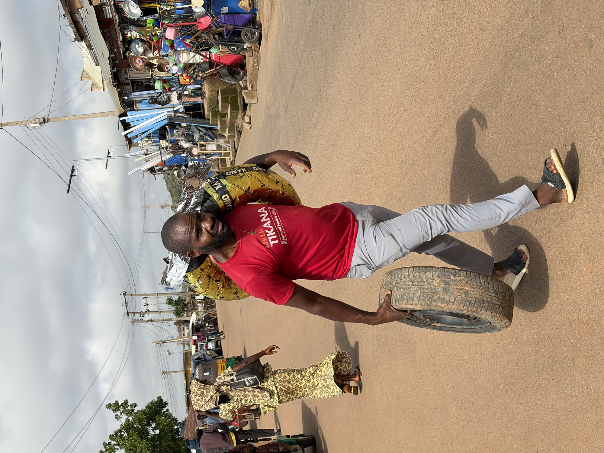 In Gura Top, Nigeria, a man in a red t-shirt and gray pants walks along a dusty road, carrying a spare tire and a large, yellow, foil-wrapped object over his shoulder. The man, with dark hair and a beard, looks towards the viewer with a serious expression. In the background, a woman in a patterned yellow dress walks away from the camera, carrying a black bag. The street is lined with market stalls selling various goods, including metal pipes, tools, and household items. Power lines crisscross the sky, and a cloudy sky suggests an overcast day. The overall scene depicts a busy marketplace environment.