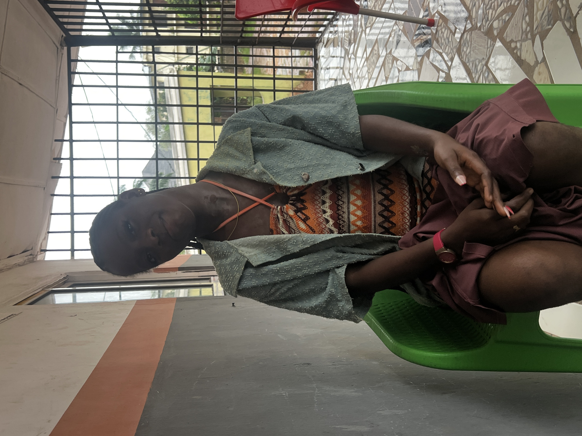 A medium shot captures a young Black woman seated in a bright green plastic chair on a tiled patio. She wears a patterned orange and brown dress and a light green collared jacket over it. A thin gold chain and an orange cord are visible around her neck. Her hands are clasped in her lap, and she wears a pink watch on her left wrist. To her right, a red chair and a partial view of a person are visible. The background features a building with an orange and grey striped facade, a window with bars, and a metal gate. The ground is covered in a mosaic of light and dark tiles. The setting appears to be an outdoor area of a residential or commercial building in Nkpor, Nigeria.