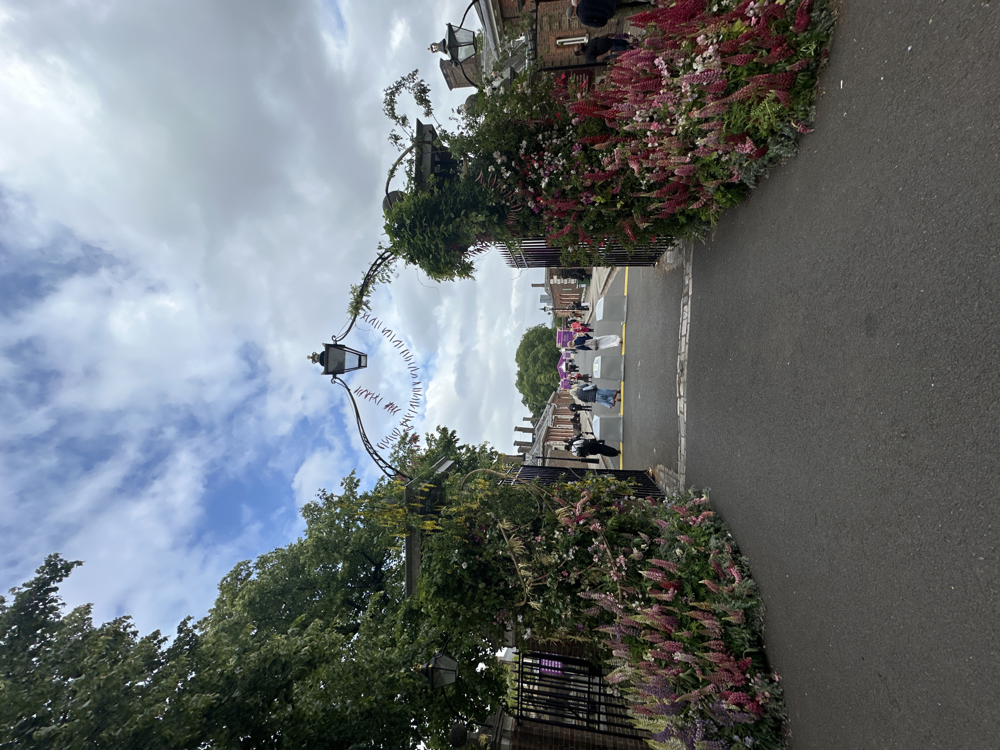 This outdoor scene in London, United Kingdom, presents a gateway. The archway is decorated with a variety of colorful flowers. Several people are walking in the street through the gateway. A black lamppost is attached to the archway. The sky is mostly cloudy. The street is paved.