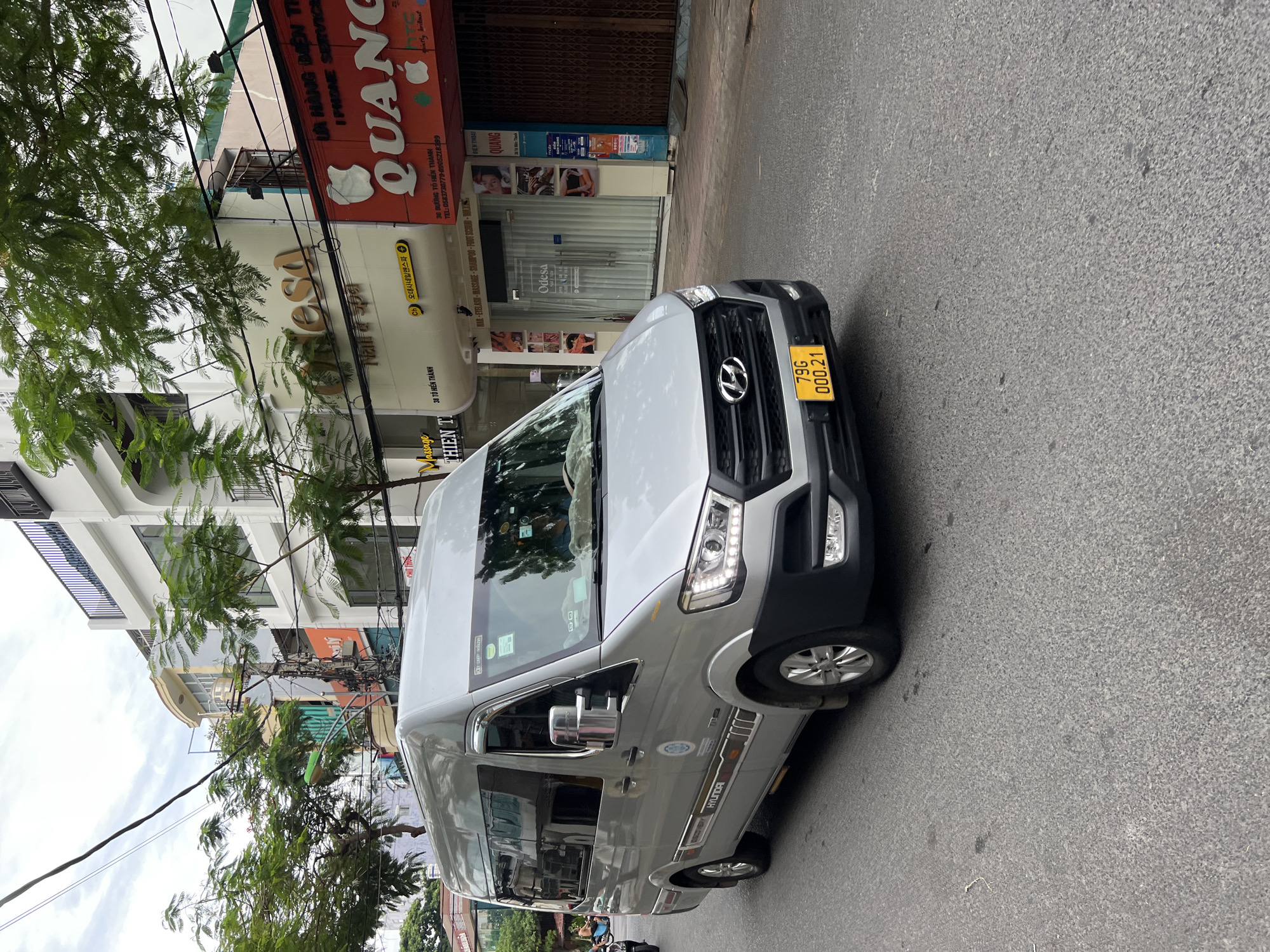 A silver Hyundai Stargazer minivan is captured in motion on a paved city street in Nha Trang, Vietnam, under an overcast sky. The minivan is the primary subject, angled towards the right side of the frame, revealing its left profile. It features tinted windows and a yellow license plate displaying "79G 000.21". The vehicle is adorned with decals on its side, including the Hyundai logo and what appears to be a circular blue emblem.
The urban environment is characterized by a row of commercial buildings lining the street. One prominent building features signage for "Odesa Nail & Spa" with Korean text above it, and another large red sign for "QUANG" with an apple logo. Further down the street, other businesses and building facades are visible, some with closed metal shutters. Trees with green foliage overhang the street and buildings, interspersed with electrical wires and poles. The road surface shows signs of wear and tear. A glimpse of another vehicle, possibly a motorcycle, is visible in the far left background.