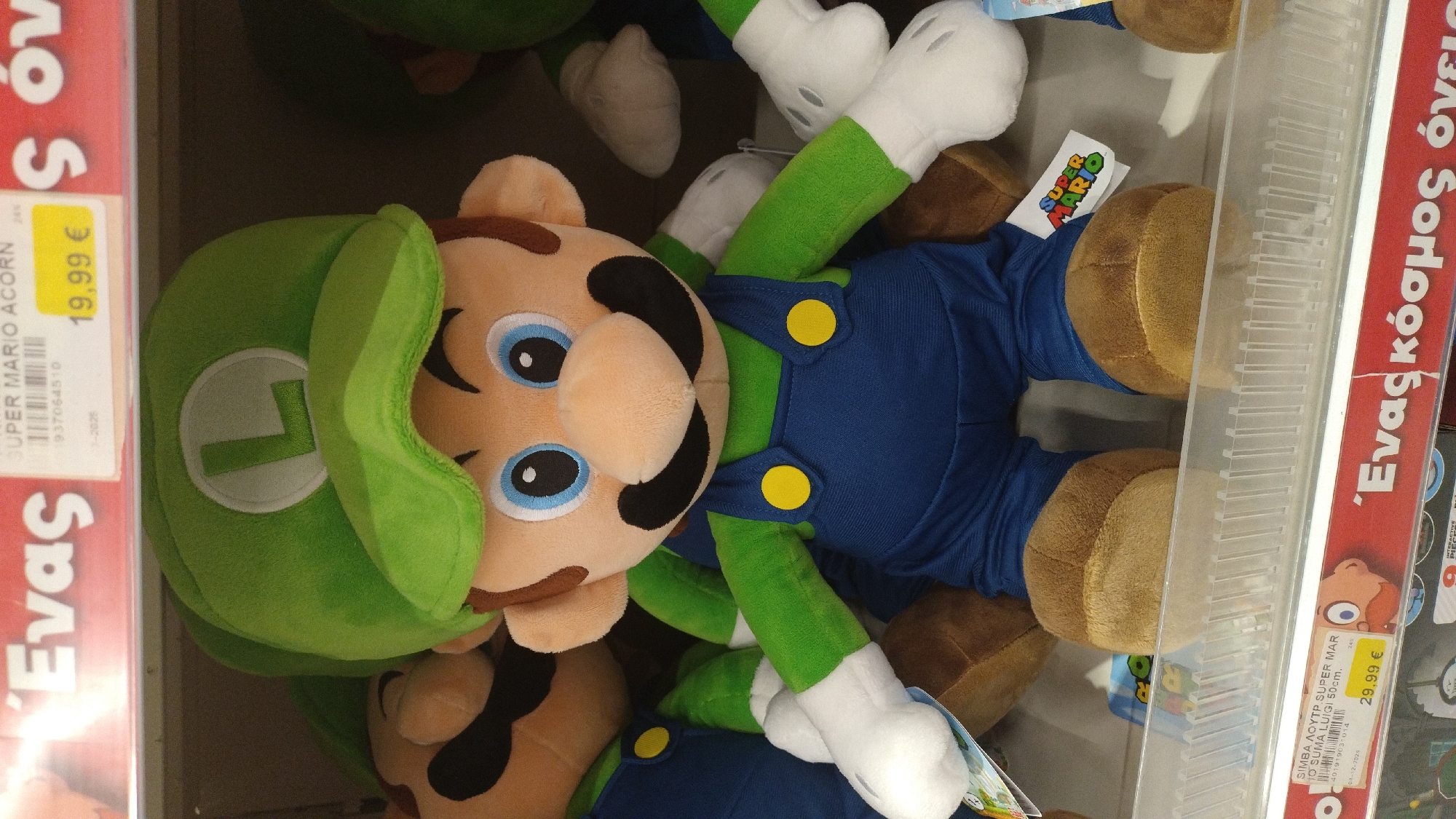 The image shows a display of Super Mario plush toys in a store, likely a toy store or department store. The central focus is a large plush toy of Luigi, recognizable by his green hat with an "L" emblem, blue overalls with yellow buttons, and green shirt. His large blue eyes, black mustache, and tan skin tone are clearly visible. To the left of Luigi, partially obscured, is a plush toy of Mario, with his red hat and blue overalls. Both toys are standing on a shelf. Above the toys, a price tag is visible, indicating "19,99 €". Further up, a red price banner displays Greek text, "Ένας" and "S όν". The background shows more merchandise on shelves and what appears to be a lighting fixture. The floor and surrounding environment are not visible, suggesting an indoor retail setting. The image does not provide information about the specific location within Pylaia Municipal Unit, Greece.