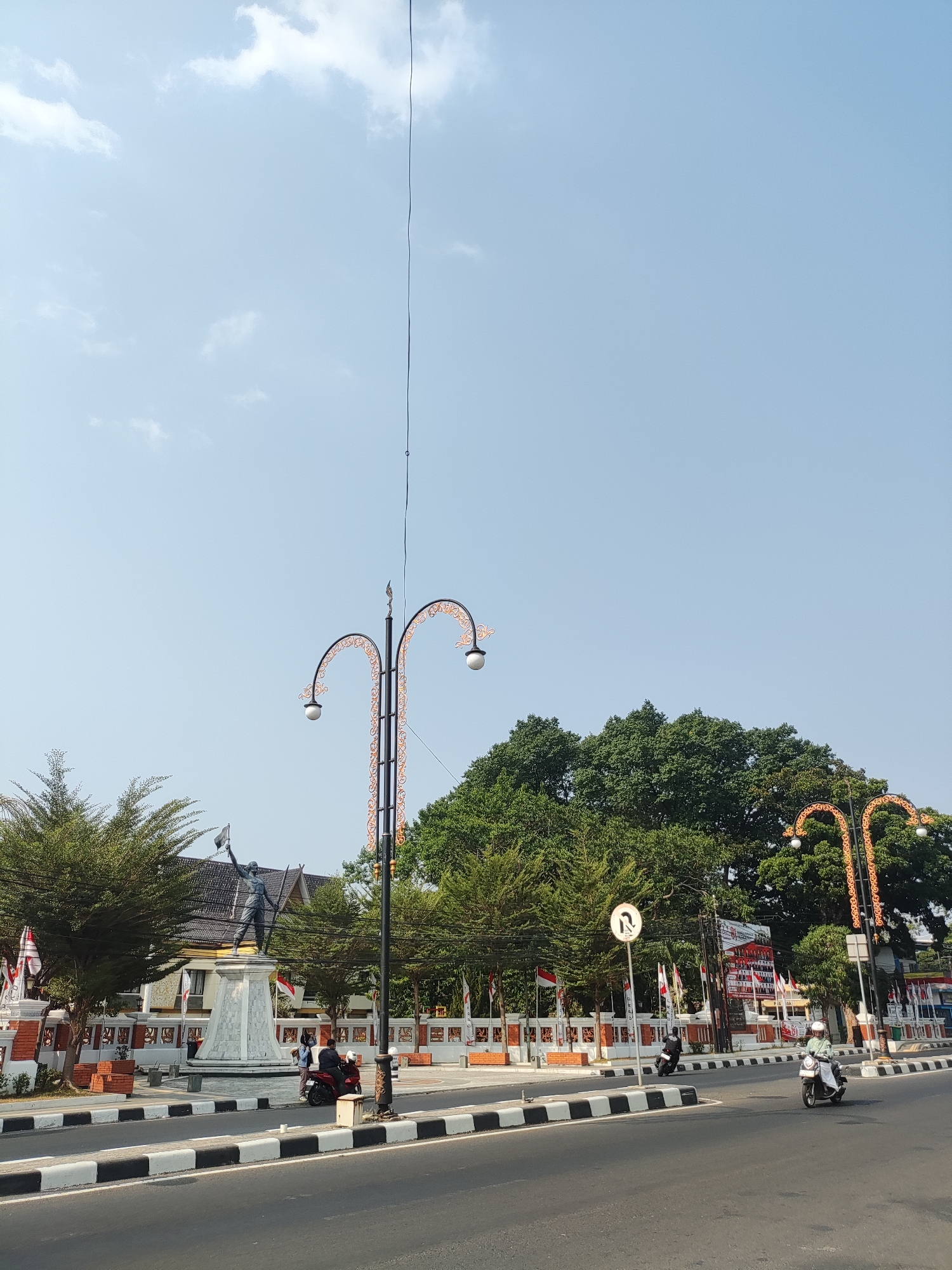 The image captures a street scene in Majalengka, Indonesia, likely a public square or intersection. In the foreground, a paved road with black and white striped dividers is visible. Two people are riding motorcycles on this road; one in the right lane wearing a white helmet and a light-colored shirt, and another in the left lane, a darker silhouette.
The midground features a sidewalk area with a low decorative wall and a large statue of a figure holding a flag aloft, standing on a tall, multi-tiered pedestal. To the left and right of the statue are trees with dense green foliage. Several Indonesian flags are displayed along the wall. Two ornate streetlights with decorative curved arms and white globes are prominent, one to the left and another to the right of the statue. Behind the statue and trees, a building with a dark roof is partially visible.
The background consists of a clear, light blue sky with a few wispy white clouds. A tall, thin pole with wires extends upwards from near the streetlight on the left. Another large tree with dark green leaves dominates the upper right portion of the image. A circular traffic sign with an arrow indicating a U-turn is visible near the right streetlight. The overall atmosphere suggests a bright, sunny day.