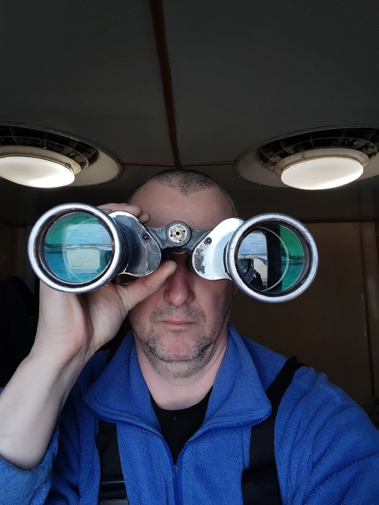 A man is holding a pair of binoculars up to his eyes. The binoculars are silver and black, with large lenses that reflect the surrounding environment. The man is wearing a blue fleece jacket and standing indoors, likely in a small room or cabin. Two round, ceiling-mounted light fixtures with metal casings are visible above him. The location is unknown.