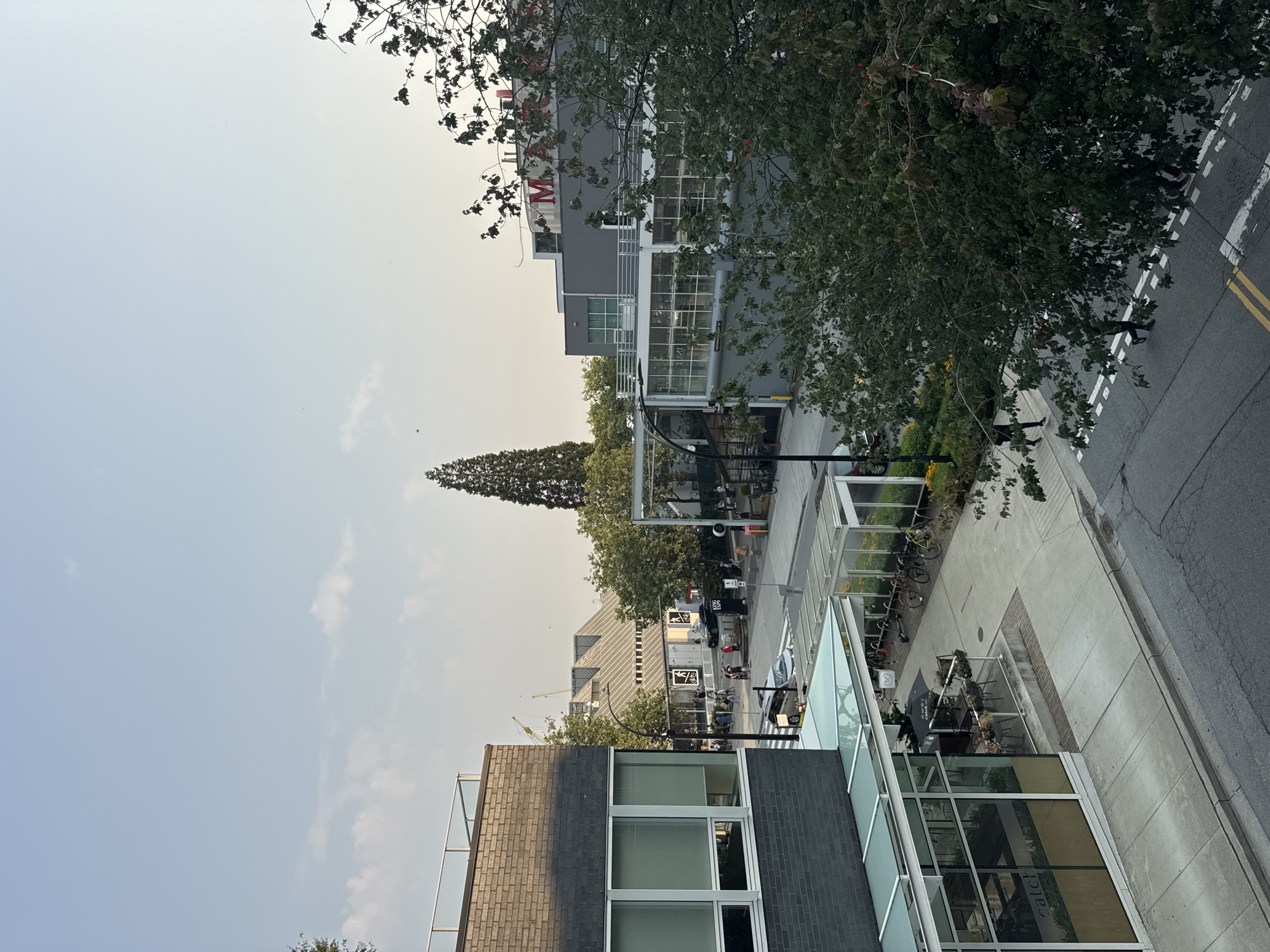 This is an eye-level, outdoor, medium shot of a street scene in North Vancouver, Canada. The scene is comprised of buildings, trees, a street, and a sidewalk. In the foreground, to the left, is a building with a glass facade and a sign that reads "catch." A sidewalk runs along the front of the building, and there are tables and chairs outside. To the right of the sidewalk, there are several bicycles parked. In the background, there are more buildings, trees, and a street. There are several people walking on the sidewalk and street. The sky is a clear blue with a few clouds.