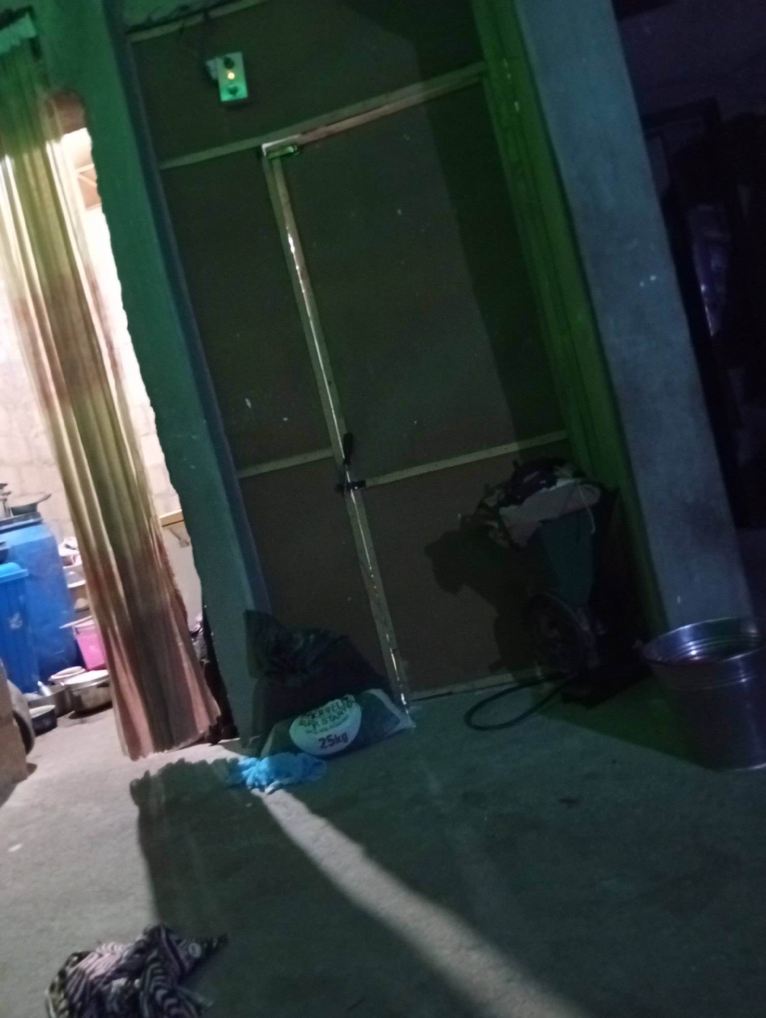 The image captures a dimly lit interior space, possibly a storage area or workshop, in Jalingo, Nigeria. A green light is visible on the upper left wall. A closed door with a metallic frame and a visible lock dominates the center of the frame. To the left, a patterned curtain hangs, partially obscuring a view into another room where blue and pink plastic containers are stacked. On the floor in front of the door, a large bag labeled "25kg" rests beside some blue fabric. Further to the right, a dark object, possibly a fan or machinery, is partially visible, and a metallic bucket containing a dark liquid is placed on the floor. The floor appears to be concrete and shows some scuff marks and shadows.