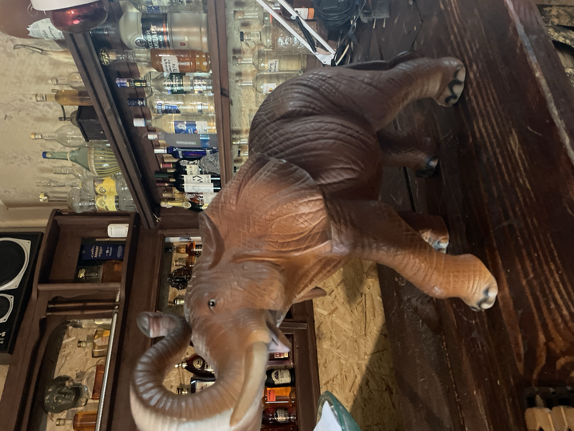 An elephant statue stands on a wooden counter in a dimly lit room, possibly a bar or shop, with shelves of alcohol bottles in the background. The statue is brown and detailed, with its trunk raised. The shelves are filled with various bottles, some clear, some dark, and some with colorful labels. The wall behind the shelves appears to be made of stone or plaster. The overall impression is one of a vintage or rustic establishment. It is impossible to determine the location in Stepanavan, Armenia from the image alone.