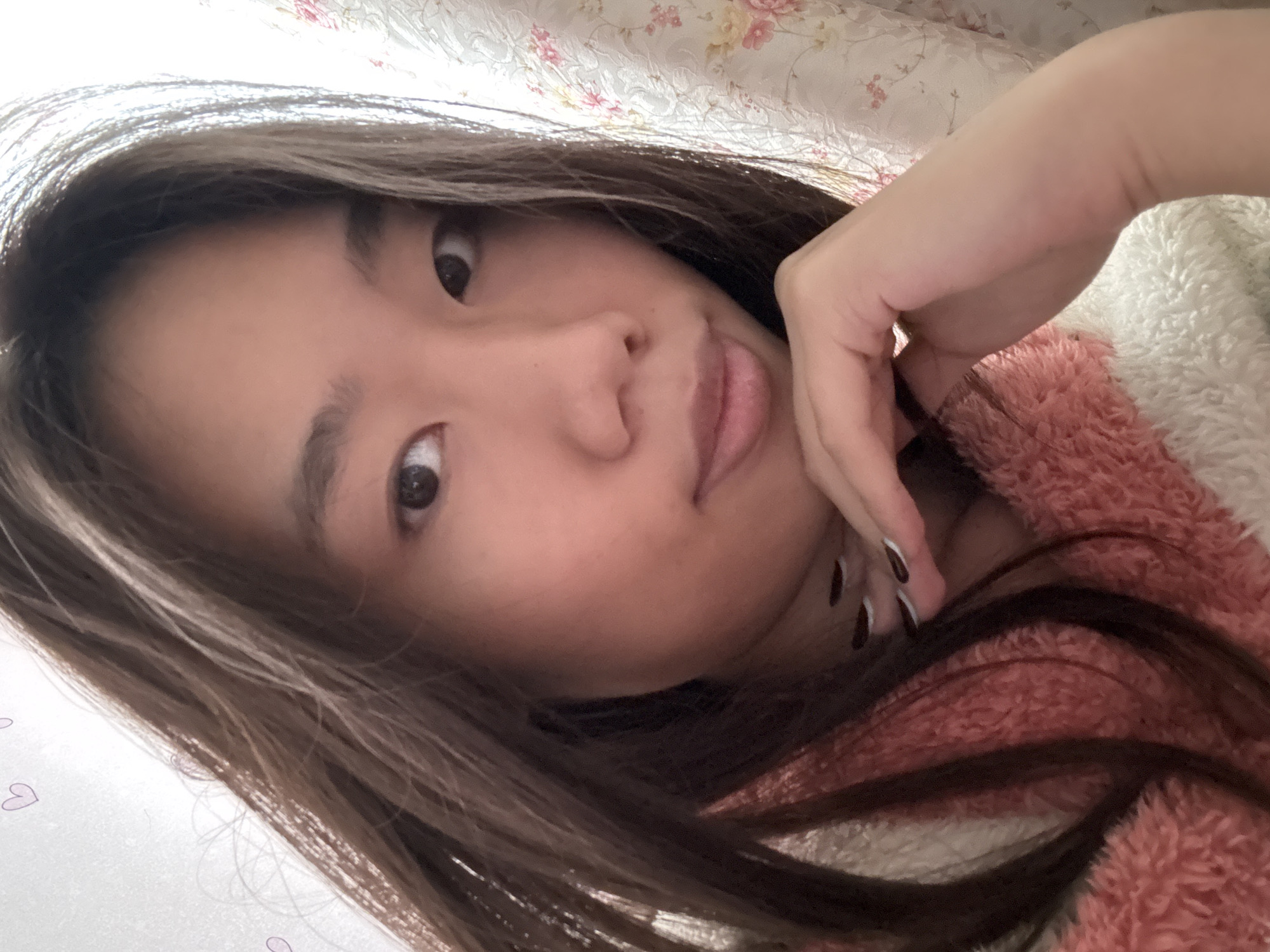 A young woman is depicted in a selfie, holding her hand to her face. She has long, dark brown hair and brown eyes. She is wearing a pink and white fuzzy jacket. The background includes a floral-patterned surface, possibly bedding. No specific information or indication of a location within Shapingba District, China, is discernible from the image.