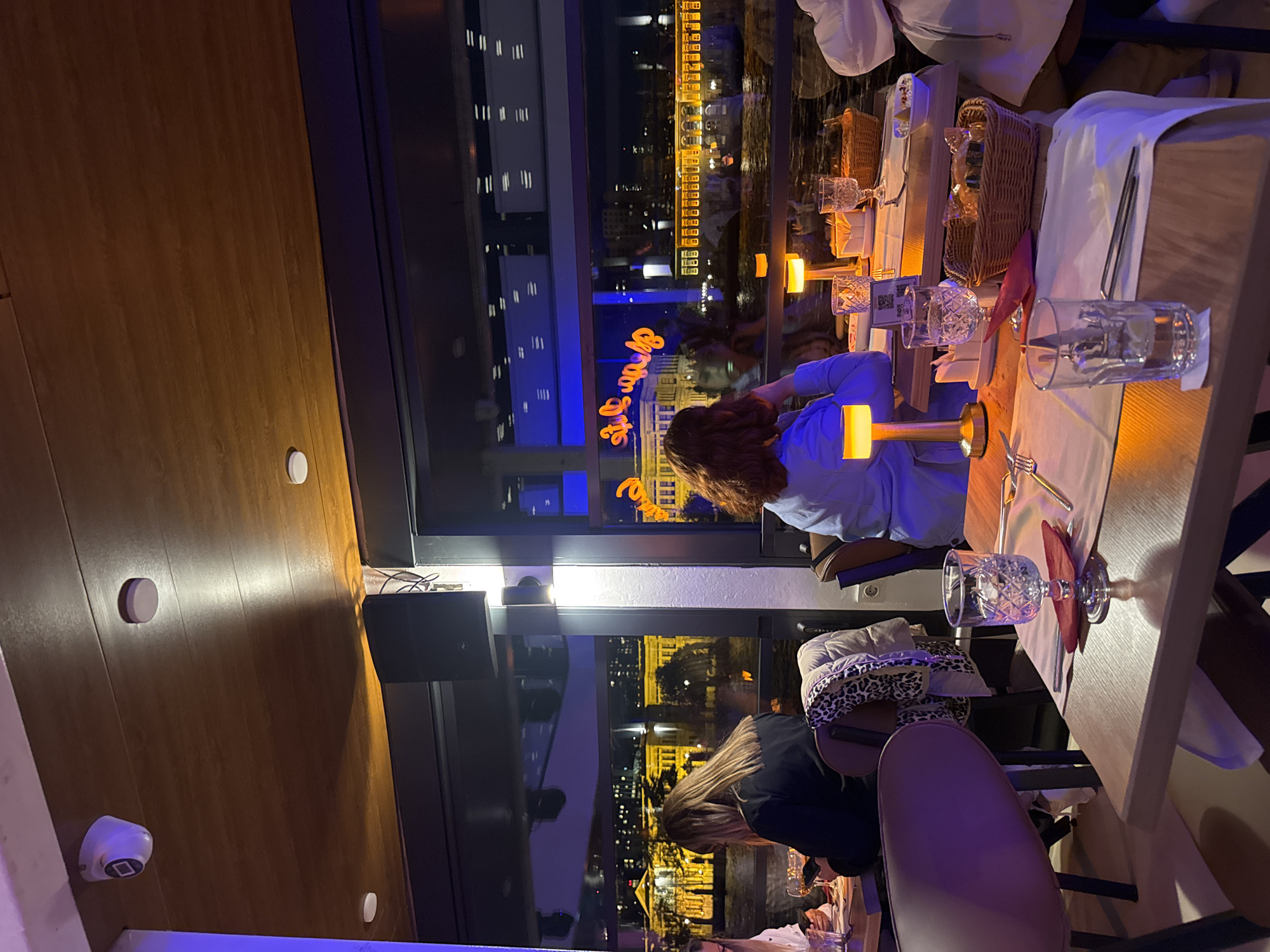 The image captures an interior view of a restaurant or cafe at night, offering a panoramic cityscape. The perspective is from inside, looking towards large windows that overlook a city with a body of water.
In the foreground and midground, multiple dining tables are visible, set with crystal glasses, white napkins, cutlery, and small, warm-toned table lamps. Several patrons are seated at these tables, seen from behind, facing the expansive window view. One person wears a light blue top, another has long, blonde hair and a dark top. On the wooden wall or ceiling, there are white circular light fixtures or sensors, a white security camera, and a black speaker. A white electrical outlet is also visible. Some tables feature wicker baskets, possibly containing bread or snacks.
The background reveals a stunning nightscape of a city, characterized by numerous illuminated buildings stretching across the horizon and reflecting in a dark body of water, likely the Bosphorus. The sky is dark, indicating evening or night.
A notable detail is a vibrant orange neon sign, reflected prominently on the window, which clearly reads "It's Demands" in a stylish cursive font. Another, smaller reflection of text is present but illegible. A small, white QR code sticker is also visible on one of the table lamps. The overall atmosphere appears elegant and relaxed, with guests enjoying the meal and the illuminated urban scenery.