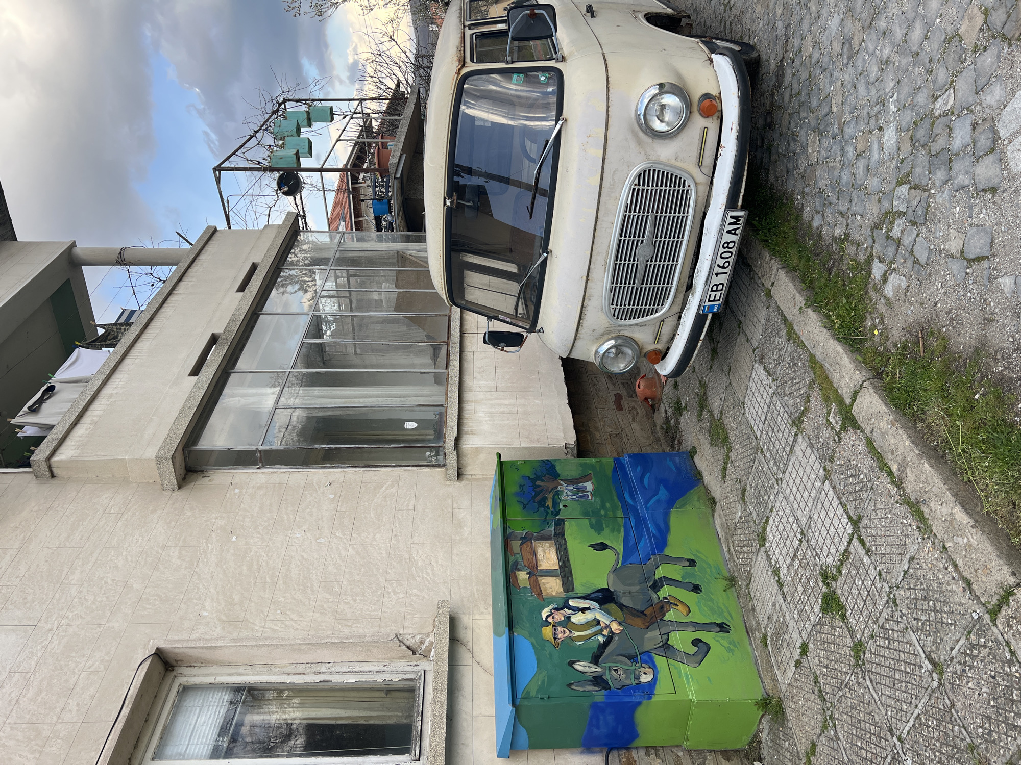 The image shows a light-colored, vintage van parked on a cobblestone street in Dryanovo, Bulgaria. The van's license plate reads "EB 1608 AM." Next to the van, against the side of a light-colored building with multiple windows, is a green utility box painted with an image of two men riding a donkey. The sky is partially cloudy. A metal structure for hanging plants or clothes is visible above the building.