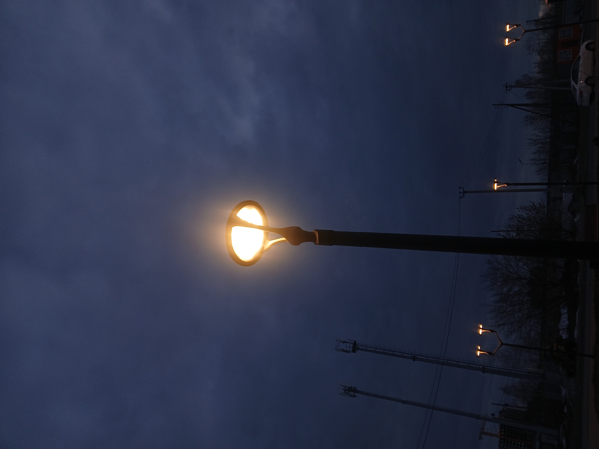 The image shows a nighttime scene with a street light in Chaikovsky, Russia. The sky is a dark blue with visible cloud patterns. The street light is prominently featured in the foreground, with a bright, round light fixture atop a black pole. Other streetlights can be seen in the distance along a road with bare trees and utility poles. A white car is parked on the right side of the road. In the lower right corner, cranes and construction are visible.