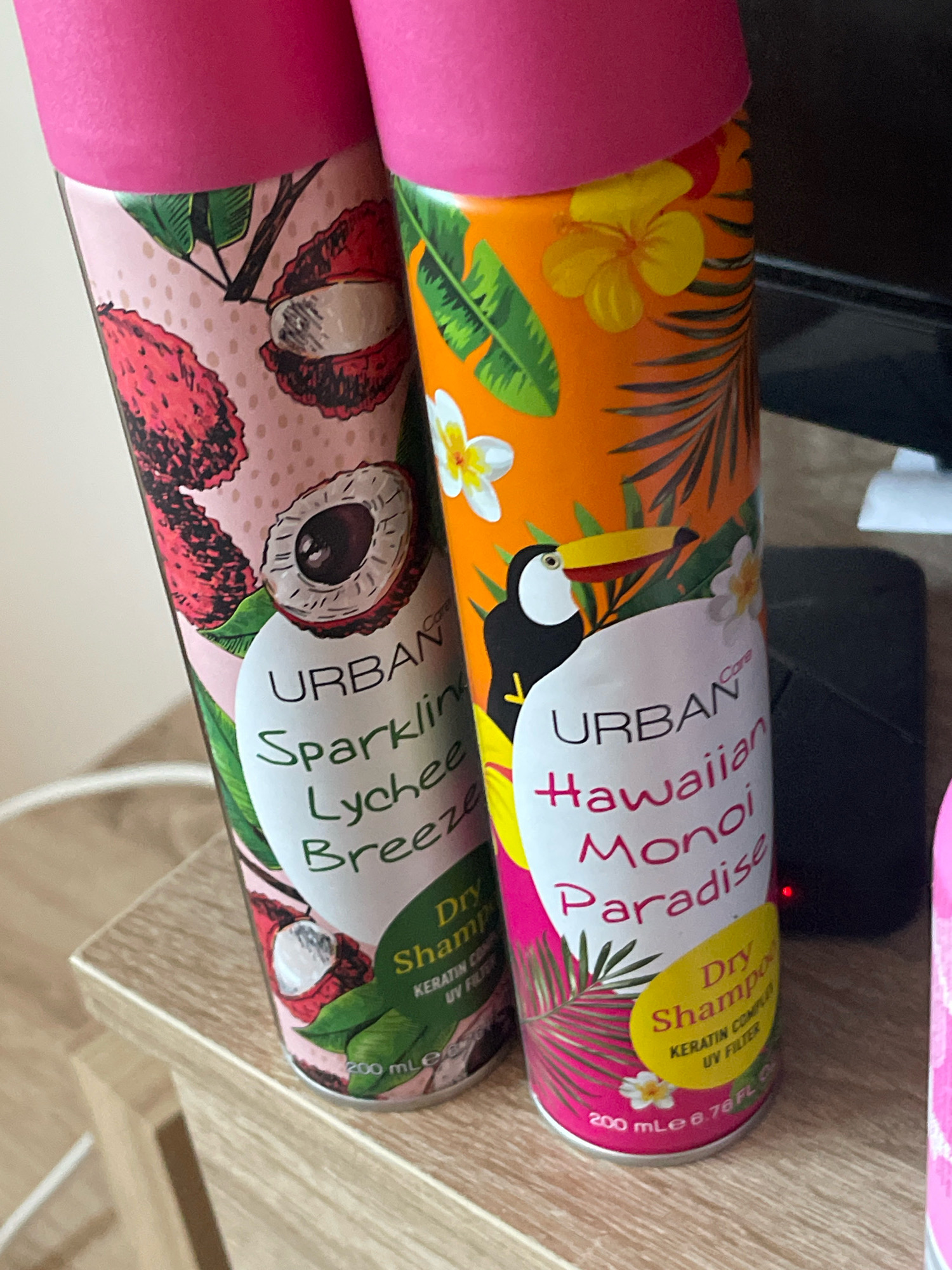 The image displays two aerosol cans of Urban Care dry shampoo positioned on a light-colored wooden surface. The can on the left features a pink lid and a design with lychee fruits and green leaves on a pale pink background. It reads "URBAN Sparking Lychee Breeze Dry Shampoo". The can on the right has a pink lid and an orange background with tropical leaves, flowers, and a toucan. It reads "URBAN Hawaiian Monoi Paradise Dry Shampoo". The cans are standing upright, and a portion of a third pink can is visible on the far right edge of the image. The lighting suggests an indoor setting. The specific location of Prizren, Kosovo, cannot be determined from the visual information provided in the image.