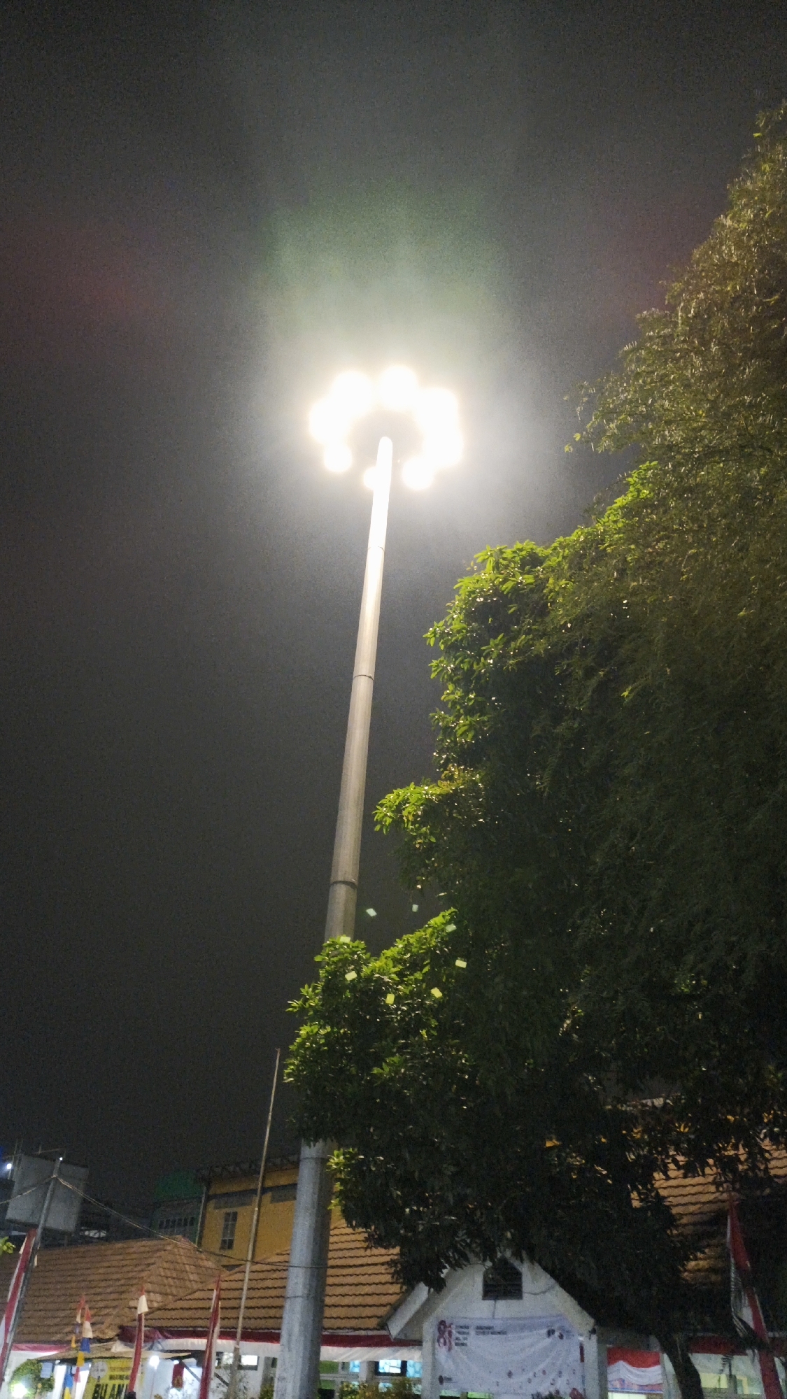 A tall, slender lamppost with multiple lights at its apex illuminates a dark night sky in South Jakarta, Indonesia. The lights cast a bright, somewhat diffused glow, creating a halo effect. To the right of the lamppost, a dense, leafy green tree obscures a portion of the scene. Below the tree, the tiled roofs of several buildings are visible, with some walls painted yellow and white. A white banner with some text, possibly a poster or advertisement, is stretched across the front of one of the white buildings. Red and white flags are also visible, suggesting a possible event or national celebration. The overall atmosphere is that of an urban environment at night, with artificial light dominating the scene.