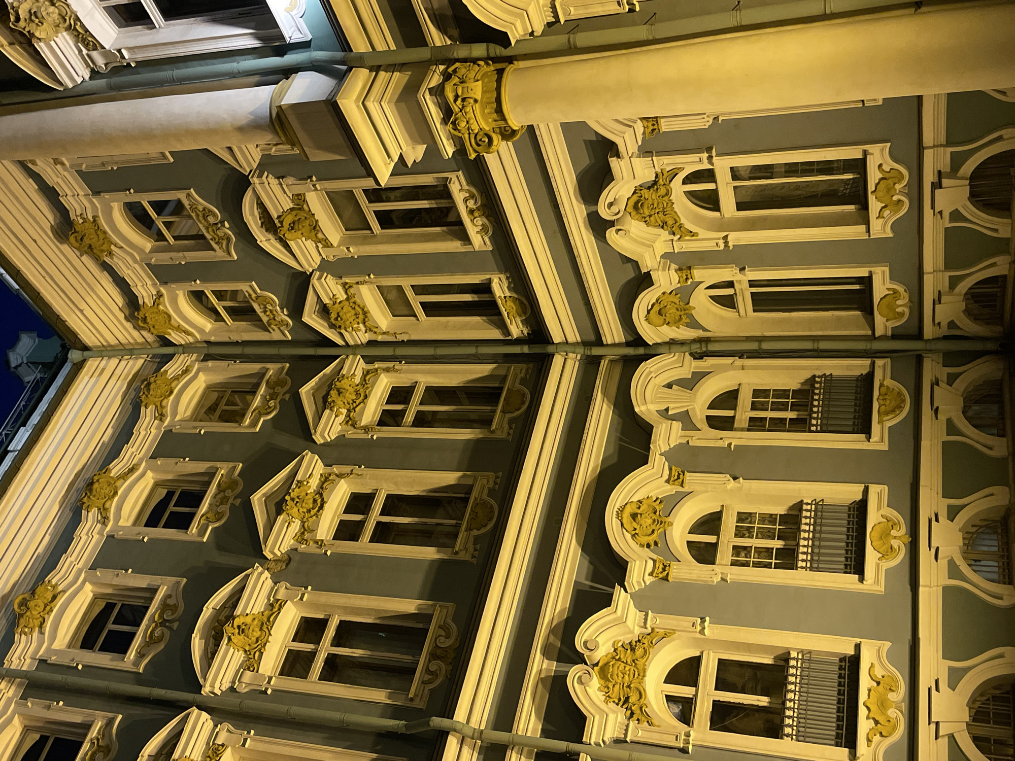 The image captures a detailed, upwards-looking view of an elaborately decorated building facade at night, likely in Saint Petersburg, Russia, as suggested by the architectural style. The scene is dominated by a repeated pattern of windows set into a blue-grey wall, richly illuminated by warm yellow light.
The building exhibits characteristics of Baroque or Rococo architecture, featuring highly ornate, golden-colored stucco decorations around the windows and on the cornices and pilasters. Some windows are rectangular with simple frames, while others are arched at the top, often featuring decorative elements resembling shells, masks, or floral motifs. Many windows have a grid-like pattern of bars, particularly those on the right side of the image.
Prominent architectural features include large, fluted columns or pilasters with ornate capitals that protrude from the facade, also highlighted by the golden light. Vertical decorative strips and cornices divide the building horizontally and vertically, creating a rhythmic structure. A narrow, dark drainage pipe runs down the facade, partially blending with the shadows.
The time of day is clearly nighttime, with artificial lighting casting strong highlights and deep shadows, emphasizing the three-dimensional quality of the architectural details. The overall impression is one of grandeur and historical elegance, typical of the opulent palaces and grand houses found in Saint Petersburg. No people are visible, and no text is discernible on the building. The image focuses solely on the intricate beauty and historical artistry of the architecture.