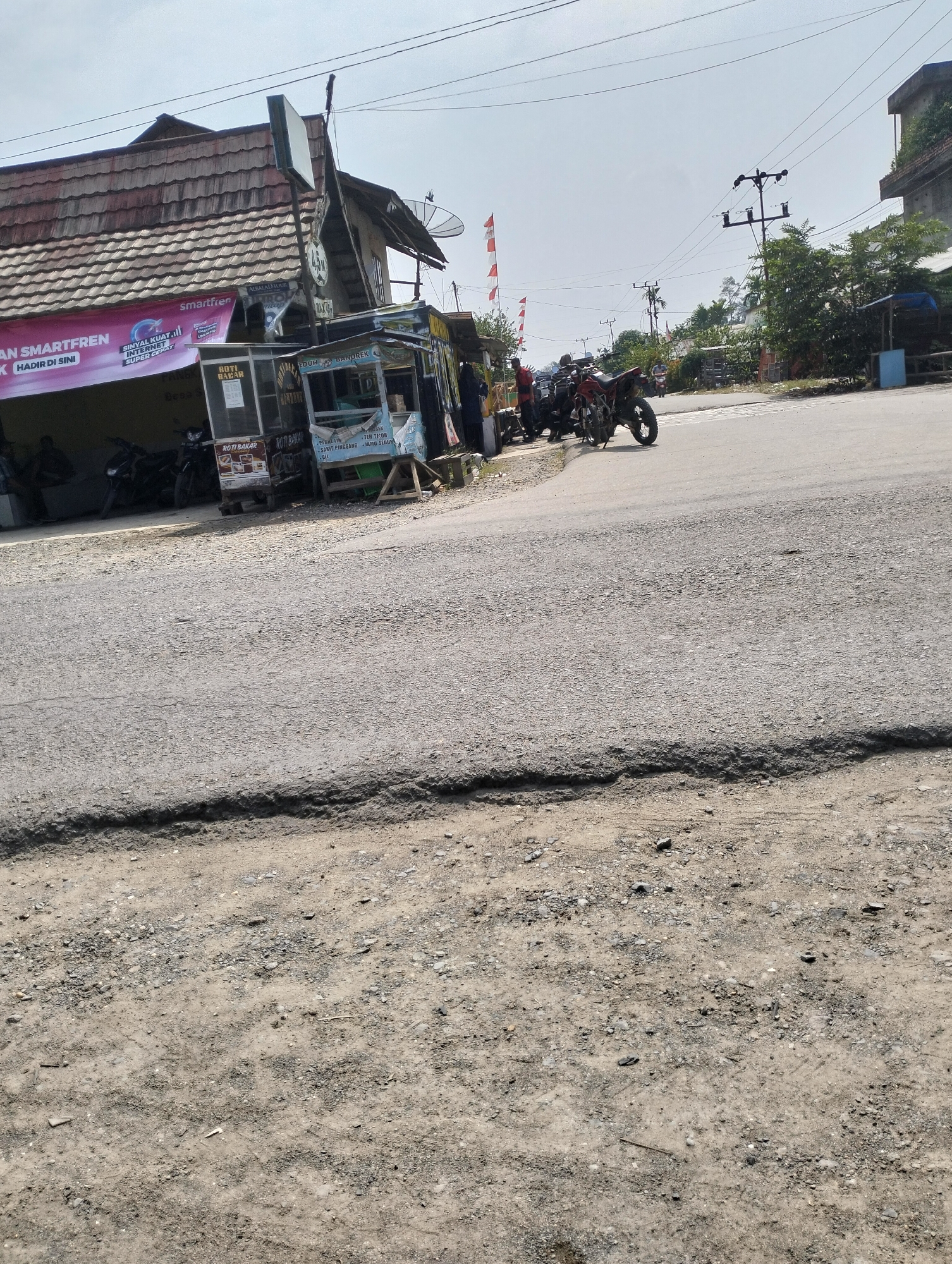 The image depicts a street scene in Pematang Lumut, Indonesia. In the foreground, a cracked asphalt road is visible, transitioning to a dirt shoulder. A small, open-air food stall is situated on the left side of the road, featuring a counter with various food items and drinks displayed. Several motorcycles are parked near the stall. Further back, a building with a reddish-brown tiled roof is visible, with signage that includes the brand "smartfren" and what appears to be promotional material. The background shows a clear sky with power lines and utility poles crisscrossing overhead. Trees and other structures line the street, suggesting a semi-urban or rural setting. A few people are present, mostly interacting with the food stall or near the parked motorcycles. Red and white Indonesian flags are displayed on flagpoles in the distance. The overall scene appears to be a snapshot of daily life on a local street.