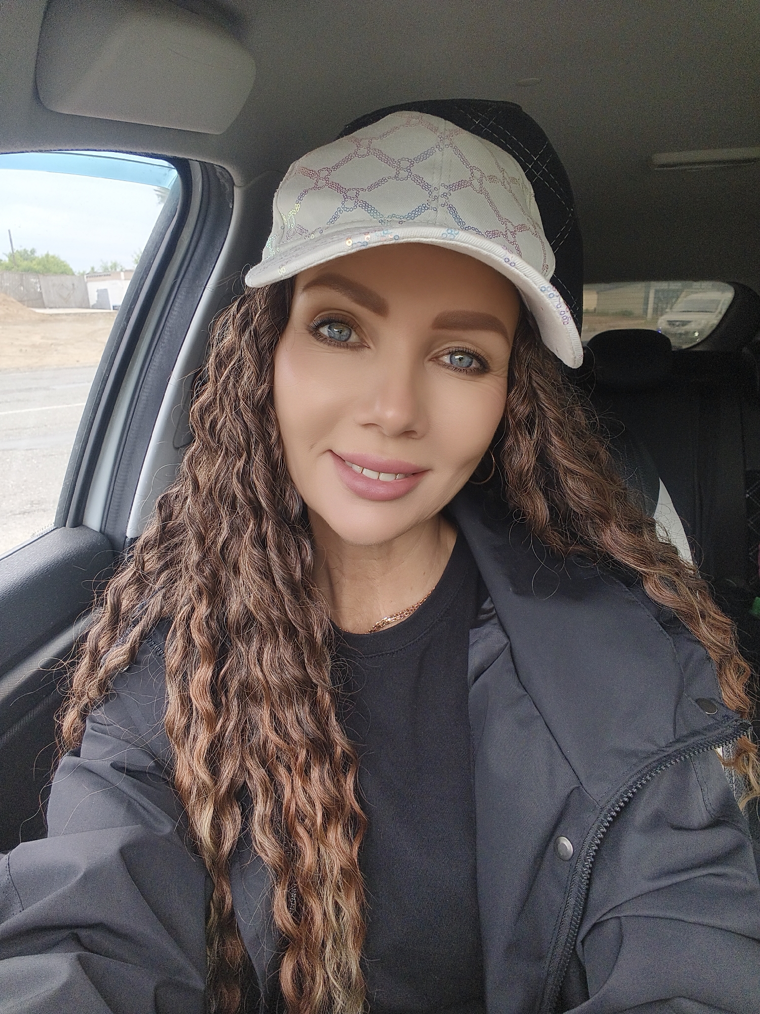 A medium shot captures a woman sitting in what appears to be the driver's seat of a car, posing for the camera. She has long, wavy or curly brown hair with lighter highlights, blue eyes, and is smiling softly. She is wearing a white baseball cap with an iridescent, possibly sequined or embroidered, pattern, a black t-shirt, and a black zippered jacket with snap buttons. A thin gold chain necklace is visible around her neck.
The interior of the car is visible, including a grey headliner, a grey sun visor, and a dark grey door panel. A blue-tinted window deflector is attached to the driver's side window. Through the window, an outdoor scene is visible with dry, light-colored ground, some low vegetation, and what appears to be concrete or stone structures in the distance, suggesting an open or possibly undeveloped area. In the background, visible through the rear window, parts of a black car seat with white accents and a partial view of another white vehicle can be seen. The overall lighting suggests it is daytime, with clear weather. There is no visible text in the image. The woman appears pleasant and calm.