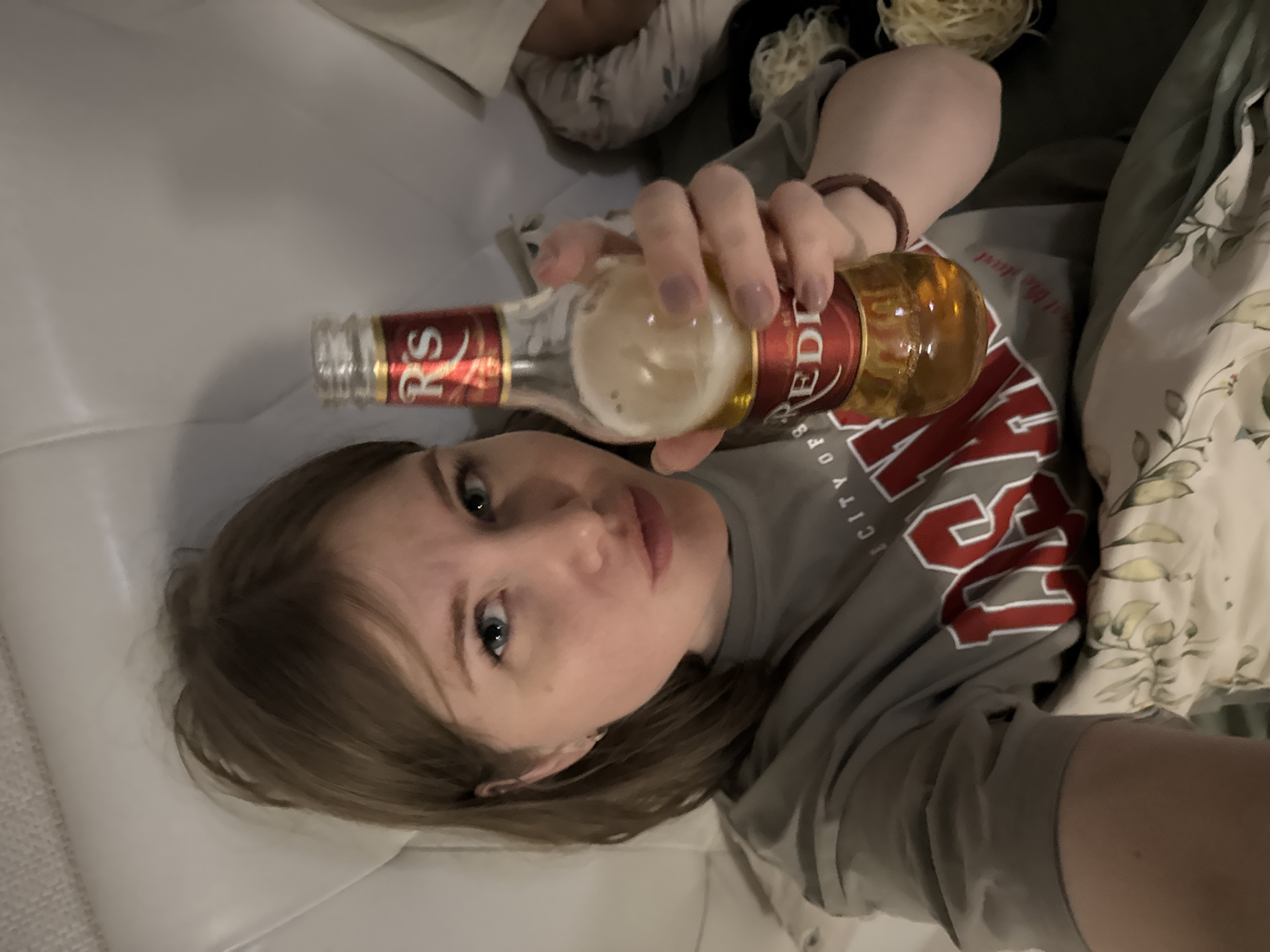 A selfie-style portrait shows a young woman lying down indoors, likely on a bed or sofa. The image is oriented vertically, with the woman's head in the lower left portion of the frame. She has light brown hair, fair skin, and blue eyes, looking directly towards the camera with a neutral expression.
She holds a clear glass bottle in her right hand, which contains a light amber liquid and has some foam near the opening, suggesting it might be an alcoholic beverage like beer or cider. The bottle features a red and gold label with visible text including "RS" near the neck and "REDDIE" prominently on the main body. Below "REDDIE," the words "CITY OF RS" can be seen. The woman wears a grey t-shirt, also featuring red and white lettering. Partial text "CITY O" and a large red letter, possibly "S" or "L," are visible on the shirt, correlating with the bottle's branding ("CITY OF RS"). A simple brown bracelet adorns her right wrist.
The woman is surrounded by bedding. A light-colored blanket or duvet with a subtle floral or leafy pattern is visible on her right, while white bedding or a pillow is beneath and to her left. In the upper left background, part of another person's leg or body is partially visible under white covers. The upper right background shows a small amount of tangled, light-colored material, possibly decorative. The setting appears relaxed and informal, consistent with an indoor space, such as a bedroom in Perm, Russia. The lighting is ambient, without specific cues for the time of day.