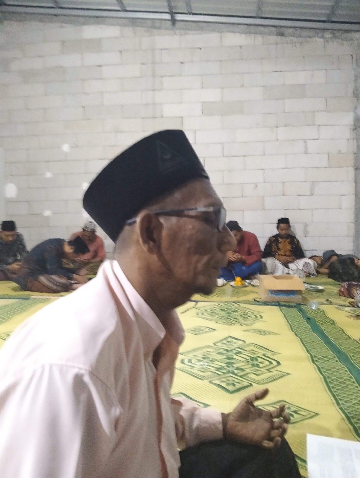 In Gedangan, Indonesia, an indoor scene depicts several men sitting on a yellow mat with green patterned borders against a light-colored brick wall. The man in the foreground, seen from the chest up on the left, wears a light pink shirt and a black peci. He is looking to the right, with his right hand resting near his lap. Behind him, other men are seated on the mat. To the left, a man in a dark shirt and peci is hunched over. Further back, a man in a reddish-orange shirt and a peci is also seated. To his right, a man in a black and gold patterned shirt and a peci is sitting upright. Beyond him, another man is lying down. In the center of the frame, a cardboard box is placed on the mat, with a small plate containing what appear to be small orange fruits. The lighting is somewhat dim, and the focus is sharper on the foreground.