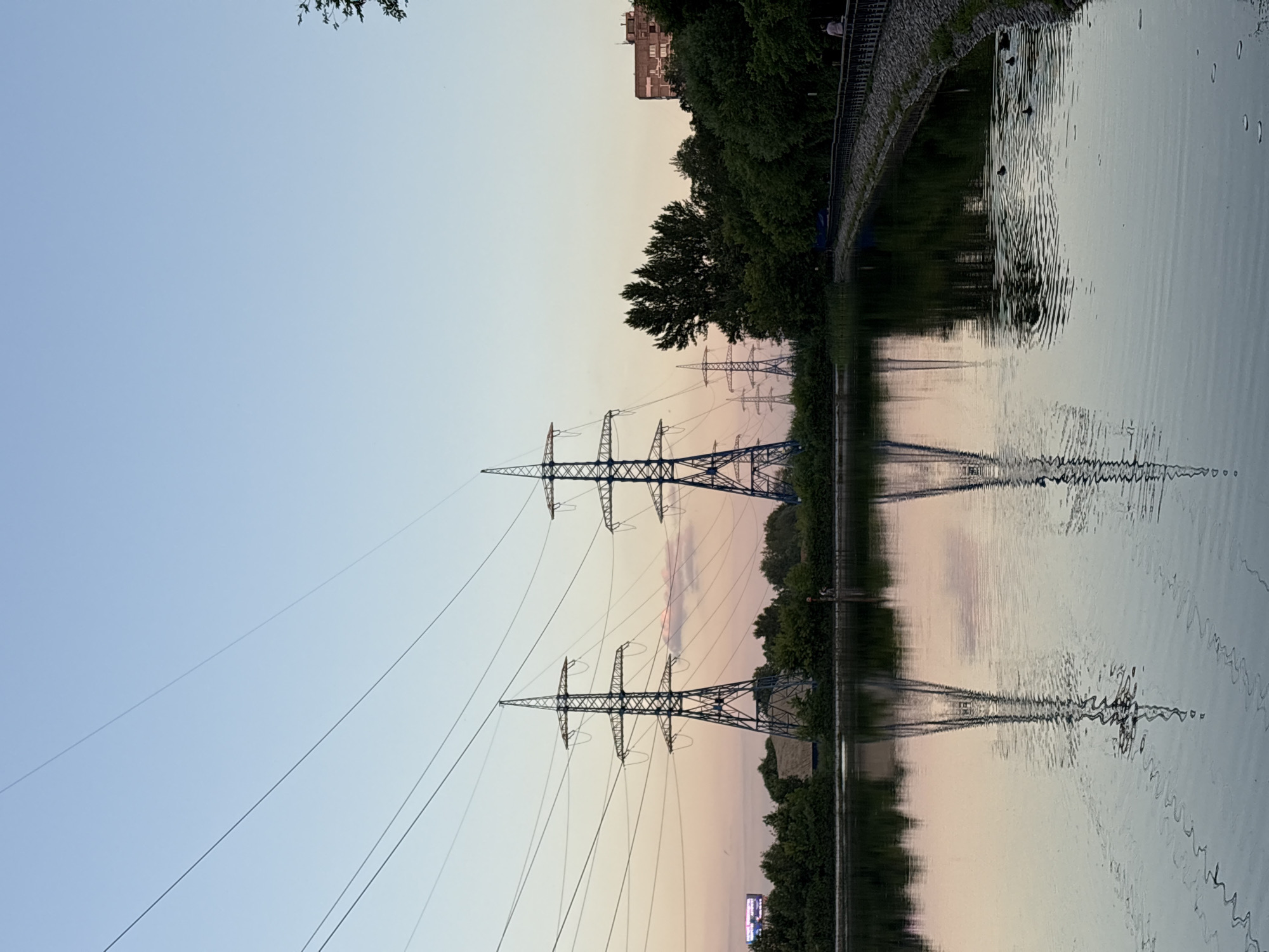 The image depicts a serene waterscape with tall electricity pylons lining the bank, reflected in the calm water. The scene appears to be set during dusk or dawn, evidenced by the soft, pastel hues in the sky and their reflection on the water's surface. The sky transitions from a pale blue at the top to a gentle pinkish-orange towards the horizon, creating a tranquil atmosphere.
Several electricity pylons, typical of high-voltage transmission lines, are prominently featured. Their intricate metal structures are silhouetted against the sky. The water is still, with subtle ripples disturbing the reflection of the pylons and the surrounding trees. Dark green foliage from trees borders the water's edge, creating a natural contrast with the man-made structures. In the background, a multi-story building can be seen above the trees, suggesting an urban or suburban setting. The water also shows a few dark, circular shapes, possibly ducks or other waterfowl, creating small disturbances on the surface.
The overall mood is peaceful and reflective, with the natural beauty of the sky and water complementing the industrial elements of the pylons. The time of day suggests a quiet moment, perhaps at the end or beginning of the day, allowing for contemplation of the interplay between nature and infrastructure. No visible text is present in the image.