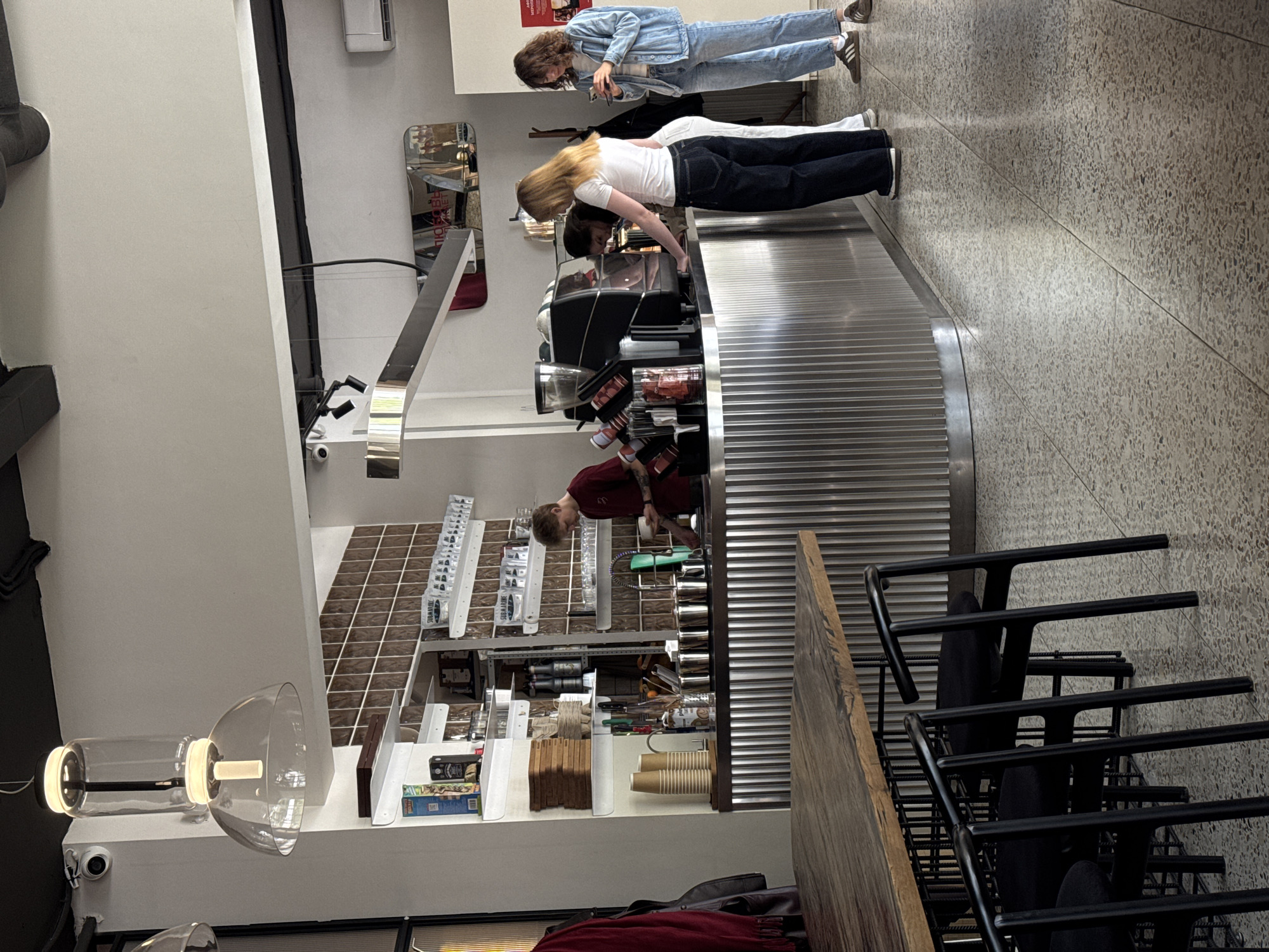 This image depicts the interior of a modern coffee shop or cafe, viewed from a slightly elevated perspective. The main focus is on a long, metallic, corrugated counter where a barista is preparing drinks for customers.
Behind the counter, a male barista with short hair and a red shirt is seen operating a professional espresso machine, with various coffee accessories and ingredients visible around him. His attention is directed towards the machine, suggesting he is actively making a beverage.
In front of the counter, two female customers are present. The customer on the left, dressed in a light blue denim jacket and jeans, is looking down at a smartphone in her hands. The customer on the right, with blonde hair, a white t-shirt, and dark pants, is leaning forward towards the counter, possibly placing an order or observing the barista at work.
The cafe's interior features a light-colored speckled or terrazzo floor and white walls. Some sections of the wall behind the counter are covered with brown square tiles, creating a decorative pattern. Shelves are visible behind the counter, holding various items such as paper cups, stirring sticks, and white containers labeled "SUGAR CUBE". Other products and packaging are also on display.
In the foreground to the right, a wooden table and several black metal chairs stacked neatly suggest a seating area within the cafe. Modern glass pendant light fixtures hang from the ceiling, illuminating the space. A small white CCTV camera is mounted on a wall near one of these lights. A reflective surface, possibly a mirror, is also visible on the wall behind the counter, reflecting parts of the interior. An air conditioning unit is mounted high on a distant wall.
The overall atmosphere appears to be that of a functional, contemporary urban cafe. The time of day is not evident, as the lighting is artificial.