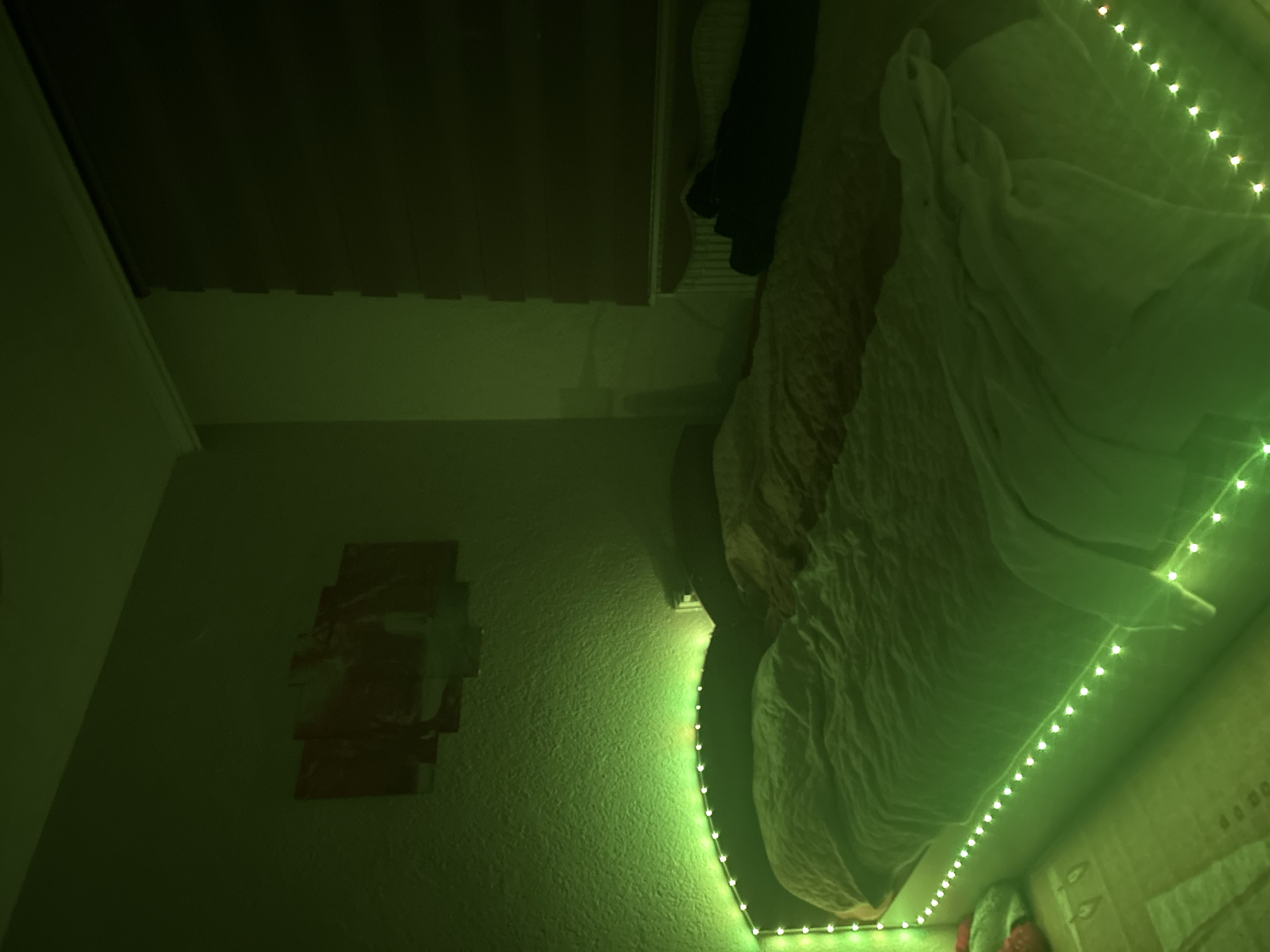 This image depicts a bedroom located in Zlatograd, Bulgaria. The room is characterized by a pervasive green ambient light, emanating from LED strip lights. These green LED strips are installed along the top curve of the bed's headboard and also along the visible side frame of the bed. The central object is a bed, covered with white bedding, beneath which an orange or brownish bedding layer is partially visible. Mounted on the textured wall above the bed's headboard is a multi-panel artwork displaying reddish tones, its specific content obscured by the green illumination. To the right of the bed, a window is fitted with a dark, possibly brown or deep red, roller blind, below which a white radiator is visible. Dark folded textiles or clothing items rest on the right side of the bed towards the foot. On the floor to the left of the bed, a colorful soft object, displaying red, green, and white hues, lies on a patterned rug. No individuals are present in the scene.