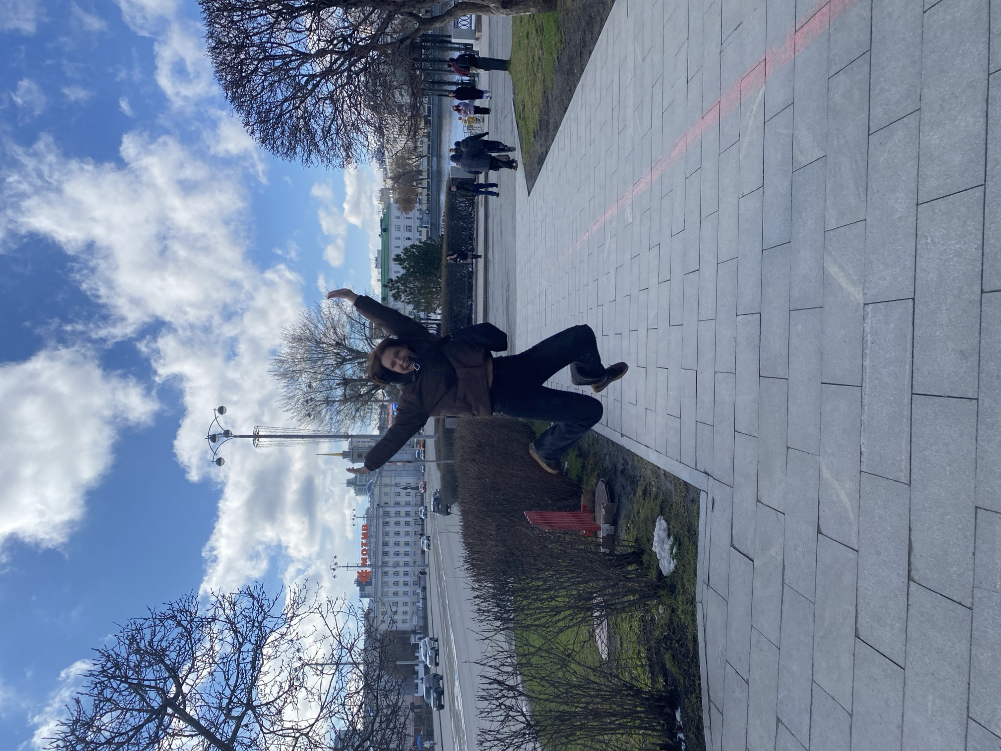 A person is in the middle of the image with both arms stretched outward. They are in front of a street with traffic and a building in the background. The building has a red sign that reads "MOTIV" in Russian. A sidewalk is on the right. People are in the background walking near water. Bare trees are on either side of the image. The sky is light blue with white clouds. The location is Yekaterinburg, Russia.