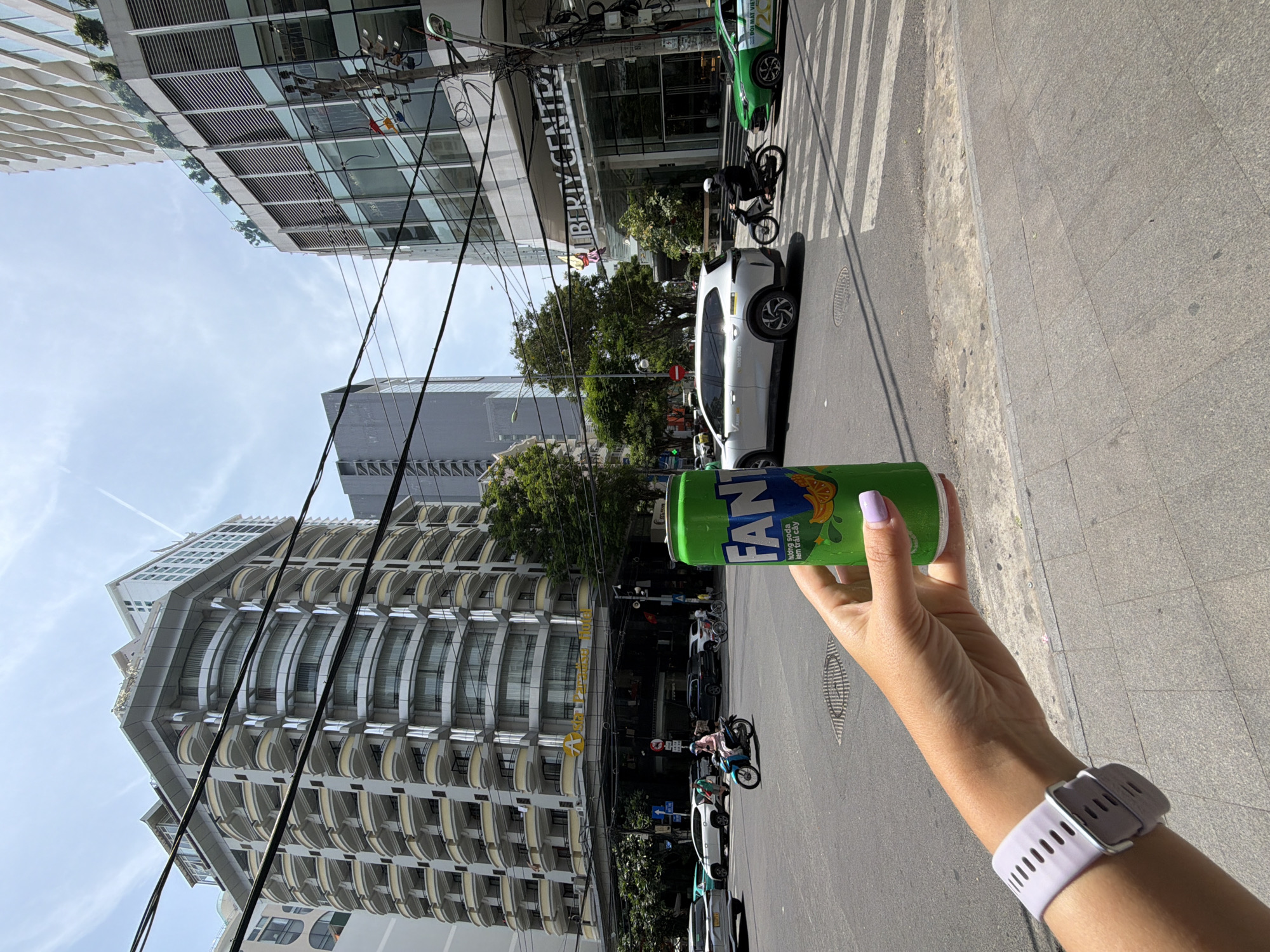 In Nha Trang Ward, Vietnam, a hand with light purple nail polish and a light purple smartwatch on its wrist is holding a green Fanta soda can. The can is positioned in the foreground, slightly to the right of the frame. In the background, a busy street intersection is visible under a bright, partly cloudy sky. Several vehicles, including a white SUV, a green taxi, and a motorcycle, are in motion on the asphalt road. Pedestrians can be seen walking on the sidewalk and crossing the street. Dominating the backdrop are tall, modern buildings with curved balconies and glass facades, one of which is labeled "LIBERTY CENTRE." Trees line the street, providing a touch of greenery amidst the urban landscape. The overall scene depicts a vibrant city environment on a clear day.