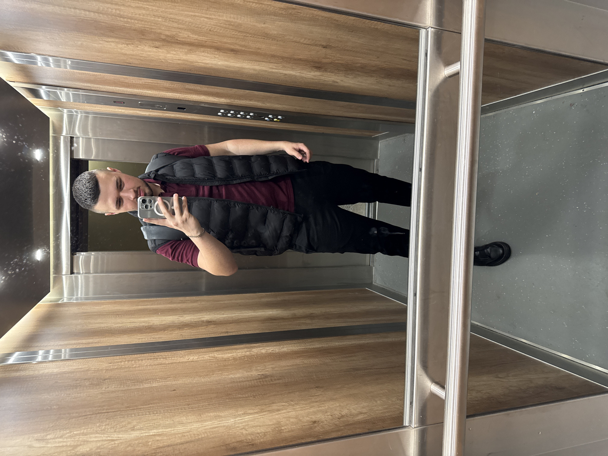 A man is standing in an elevator, taking a selfie. The elevator has wood paneling on the sides and a mirrored back wall. The floor is gray. The man is wearing a maroon shirt, black pants, a black puffer vest, and black shoes. The location is not discernible from the image.