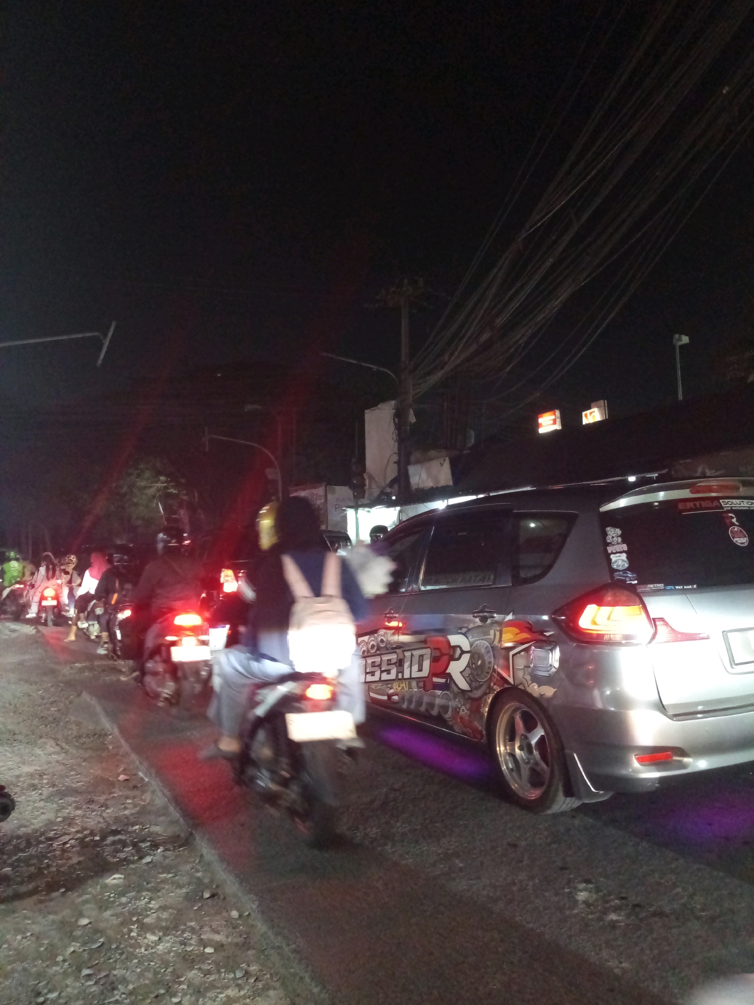 The image captures a nighttime scene of traffic on a road in Karangsatria, Indonesia. A line of motorcycles is visible in the foreground and middle ground, with riders wearing helmets and some carrying passengers. The motorcycles have illuminated taillights, indicating they are in motion or stopped. To the right, a silver car is parked or moving slowly. The car is adorned with various stickers, including one with the text "SS.ID" and a graphic of a stylized character. Purple light emanates from beneath the car, possibly from underglow lighting. In the background, buildings, streetlights, and a dense network of overhead wires are discernible against the dark sky. The road surface appears to be made of asphalt and gravel. The overall impression is of a busy urban street at night, with vehicle traffic and illuminated elements contributing to the scene.