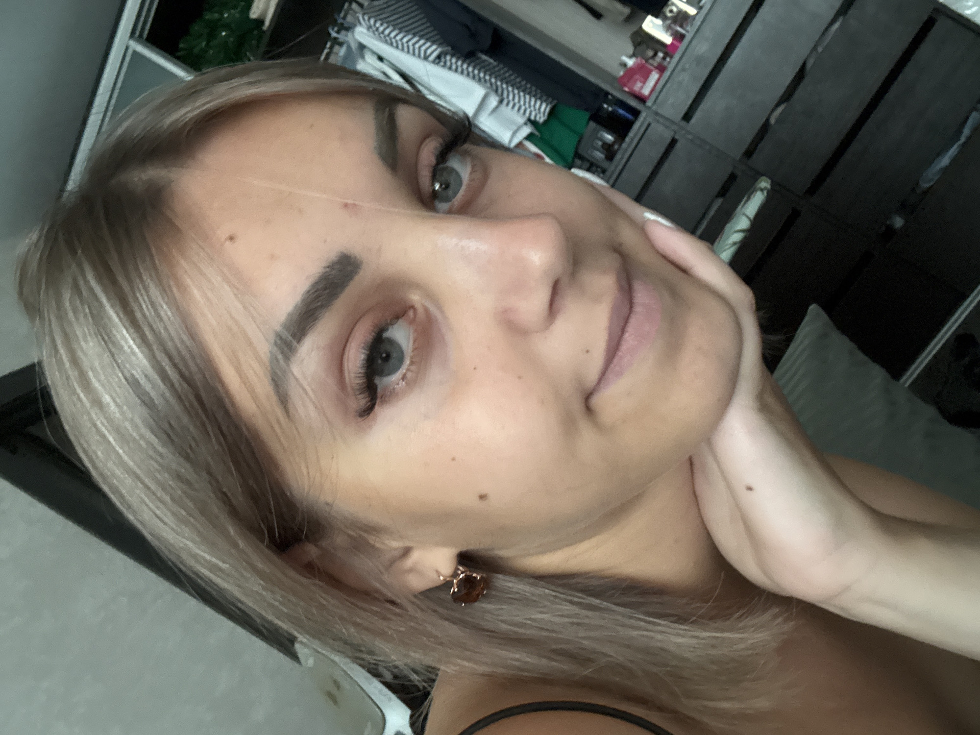 The image is a close-up, eye-level, selfie-style photograph of a young Caucasian woman with blonde hair and blue eyes. She is looking towards the camera with a slight smile, resting her chin on her hand. She is wearing makeup, including mascara and what appears to be lipstick or lip gloss. She has a heart-shaped earring in her left ear. The background is out of focus but appears to be an indoor setting, possibly a bedroom or closet, with clothing visible on hangers and drawers. There is no information in the image that would allow for identification of a specific city or location, including Yuzhno-Sakhalinsk, Russia.