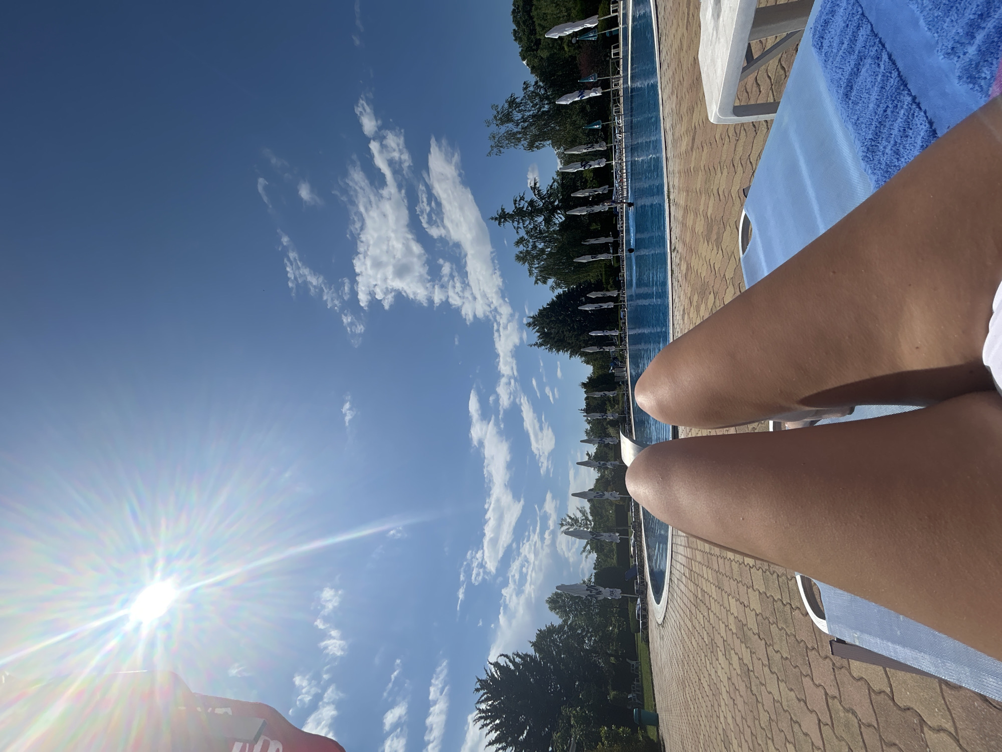 In an outdoor scene on a sunny day, a person's legs are visible in the foreground, reclining on a blue lounge chair with a blue towel. The background features a swimming pool with people swimming. Surrounding the pool area are trees, umbrellas, and lounge chairs. The sky is blue with scattered clouds, and the sun is bright. The location is identified as Unknown, Unknown.
