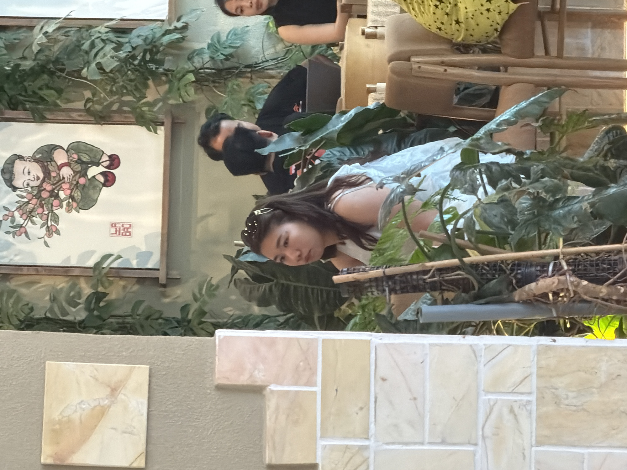 A woman wearing a white top and sunglasses on her head is seated indoors, looking to her left. She is surrounded by lush green plants. Behind her, a man in a black shirt is partially visible, with another person in a black top to his right. The background features artwork on the wall and furniture. The scene appears to be set in a cafe or restaurant in Nha Trang, Vietnam, as indicated by the presence of decorative elements often found in such establishments in the region, including the style of artwork and the prominent use of tropical plants.