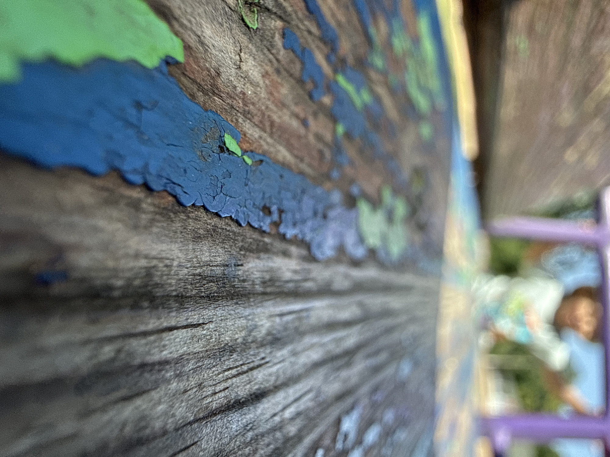 The image depicts a close-up, low-angle view of a wooden surface with peeling paint in blue and green. The wood grain is prominent, showing signs of wear. In the background, out of focus, a person appears to be on a purple structure, possibly a playground slide or swing. The setting appears to be outdoors, with greenery visible in the distance. The image does not provide specific information about its location within Piatykhatky, Ukraine.